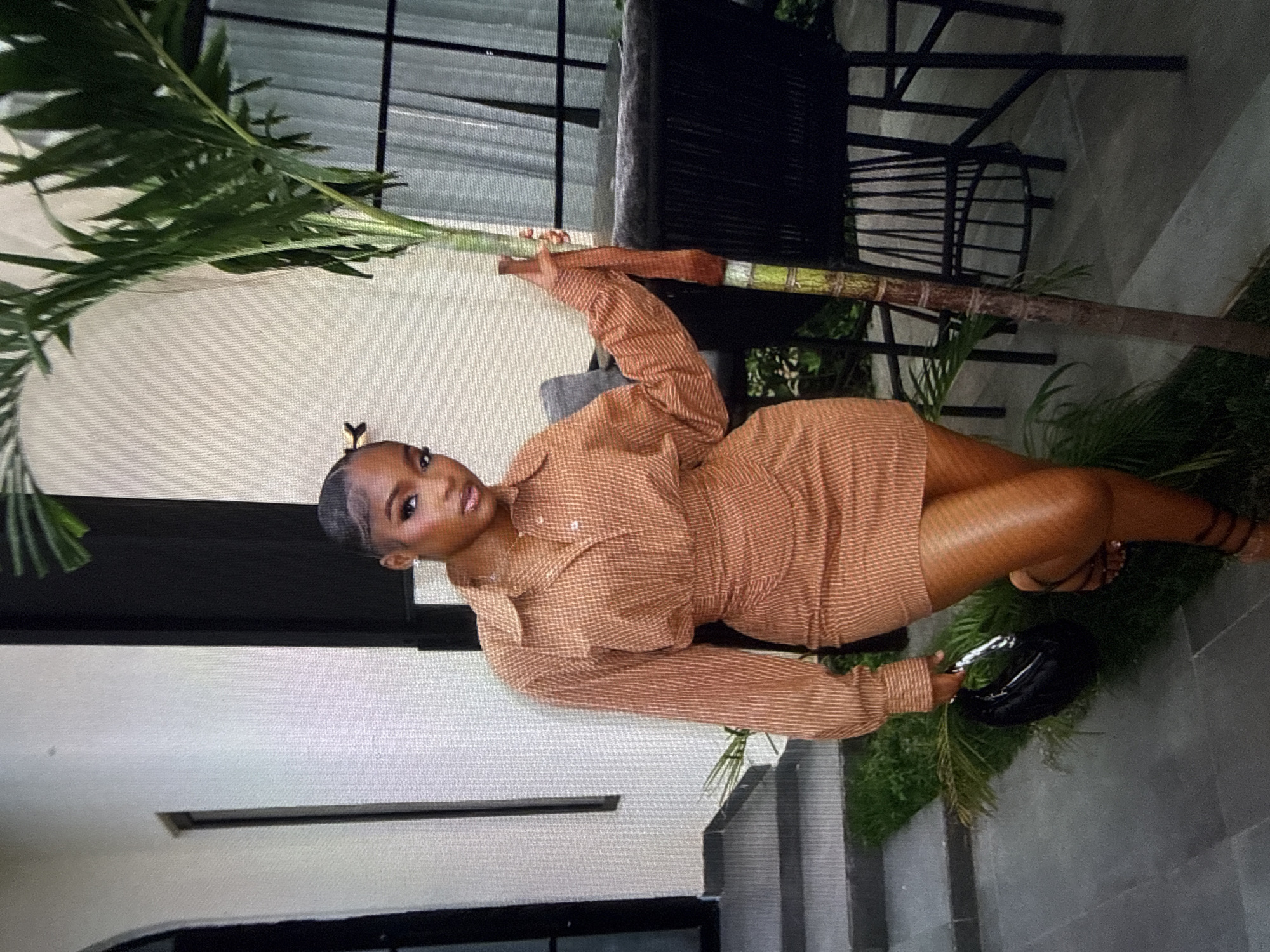 A young Black woman stands outdoors, leaning against a palm tree. She is wearing a long-sleeved, collared, rust-colored dress with a subtle checkered pattern. The dress is cinched at the waist and has a high hemline. She is holding a black, rounded handbag. Her hair is styled up, and she has on dark sandals. The background shows modern architecture with clean lines and outdoor seating. The setting appears to be a courtyard or patio area, possibly in a residential or hospitality establishment in Abuja, Nigeria, given the presence of palm trees and contemporary design elements.