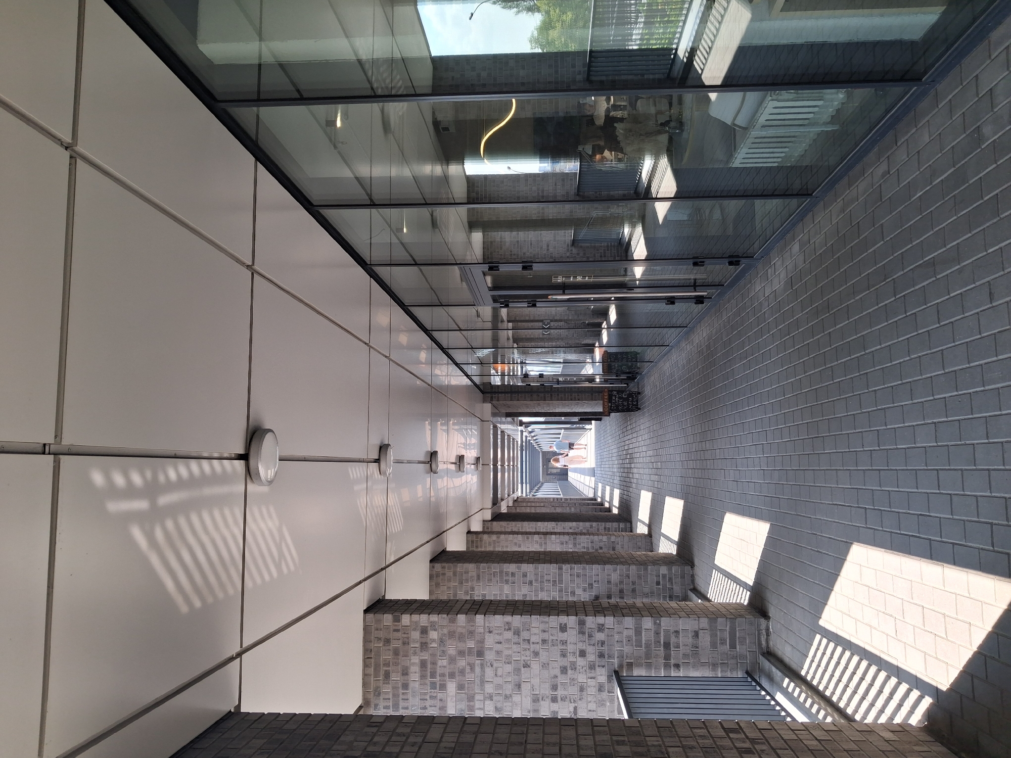 The image shows a long, covered walkway with a paved floor and a white ceiling. The walkway is lined with gray brick pillars on the left and large glass windows on the right. Sunlight casts diagonal shadows across the walkway from the pillars. Two people, a man and a woman, are walking away from the camera down the walkway. Inside the building, visible through the glass, are tables and chairs, suggesting a cafe or restaurant. The scene appears to be a modern commercial or residential building. The specific location in Novosibirsk, Russia, is not identifiable from the visual information provided.