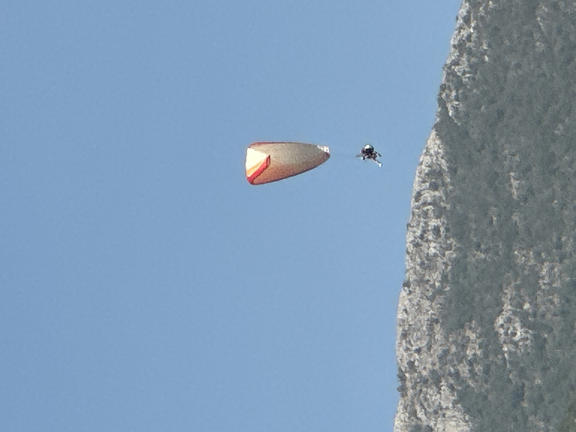 A lone paraglider is captured mid-flight against a clear blue sky, with a mountainous landscape visible in the lower half of the frame. The paraglider's wing is primarily white and orange, with a distinct teardrop shape. A person is suspended beneath the wing, appearing to be in a seated harness. The paraglider is positioned in the upper right quadrant of the image, at a considerable distance from the camera. The mountain in the foreground is covered in sparse vegetation and rocky outcroppings, appearing arid and textured. The scene is illuminated by natural daylight, suggesting an outdoor activity on a clear day. The location is identified as Boreti, Montenegro.