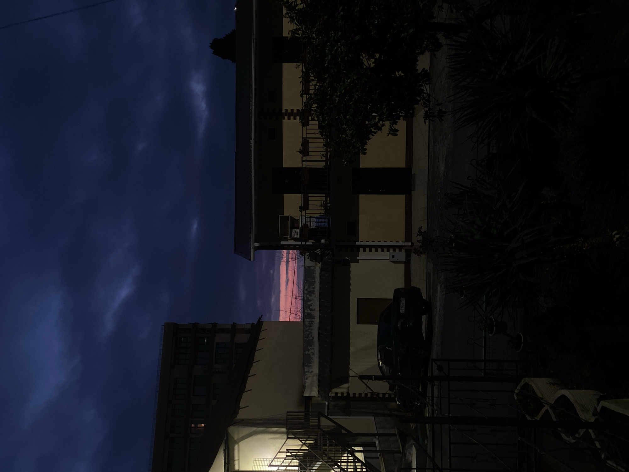 The image shows a view in the city of Sochi, Russia, at dusk. In the foreground, there are buildings with exterior staircases. A dark car is parked near one of the buildings. The sky is dark blue with hints of pink and orange in the distance between the buildings, indicating a sunset or twilight. The street is dimly lit. The focus is on the urban environment.