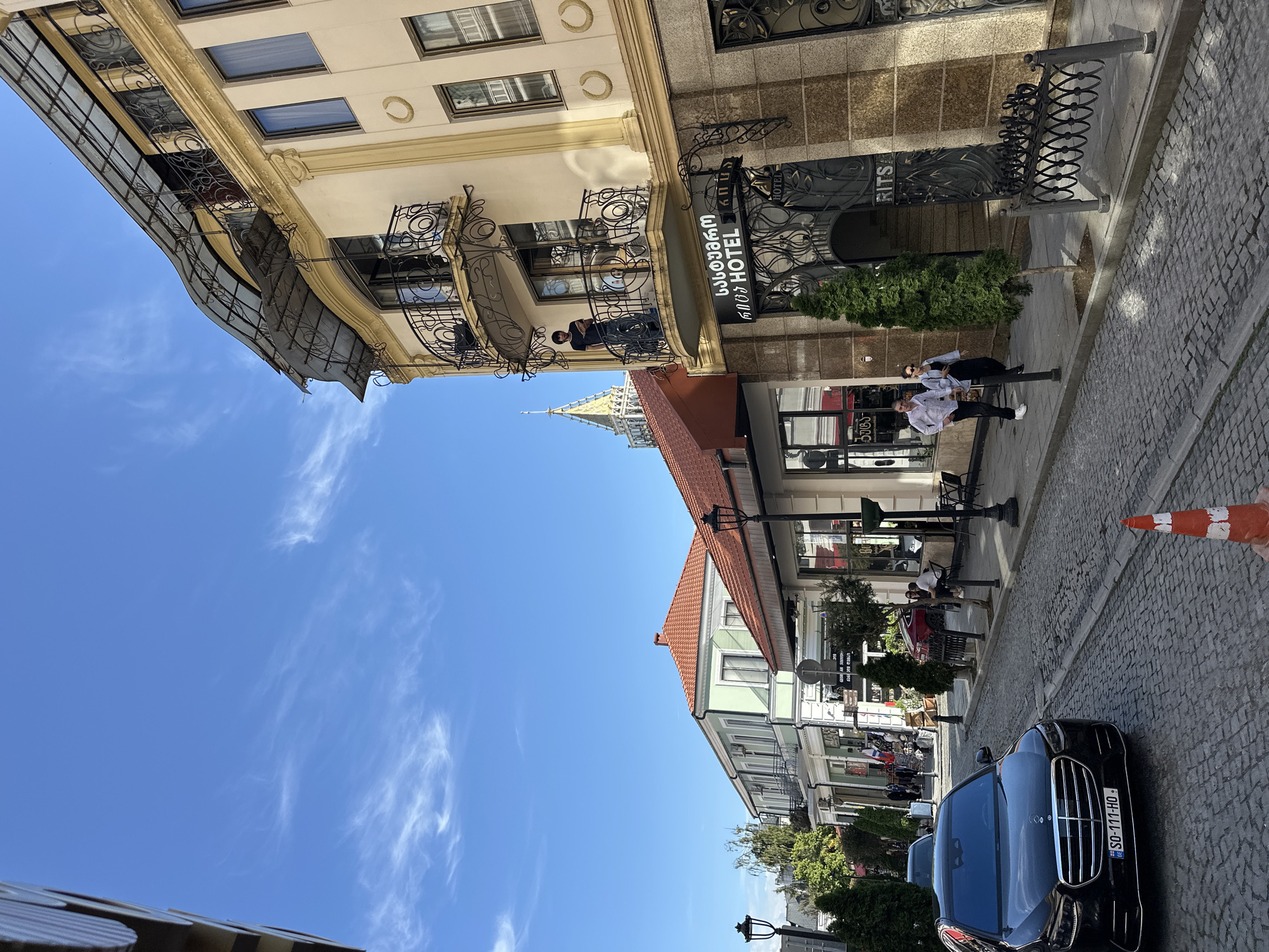 The scene captures a street in Batumi, Georgia. The sky is clear blue with wispy clouds. The street is paved with cobblestones and lined with buildings of varying architectural styles. On the left, a cream-colored building with ornate iron balconies and a gray awning is visible. A person is standing on one of the balconies. A sign for "Rits Hotel" in Georgian and English is mounted to the building. Two people stand on the sidewalk near the entrance to the hotel. A black car with a Georgian license plate (SO-111-HO) is parked on the right. A traffic cone is near the car. Further down the street, the buildings have red tile roofs and green or cream-colored facades, with lamp posts lining the sidewalk. People are seated at tables outside a cafe.