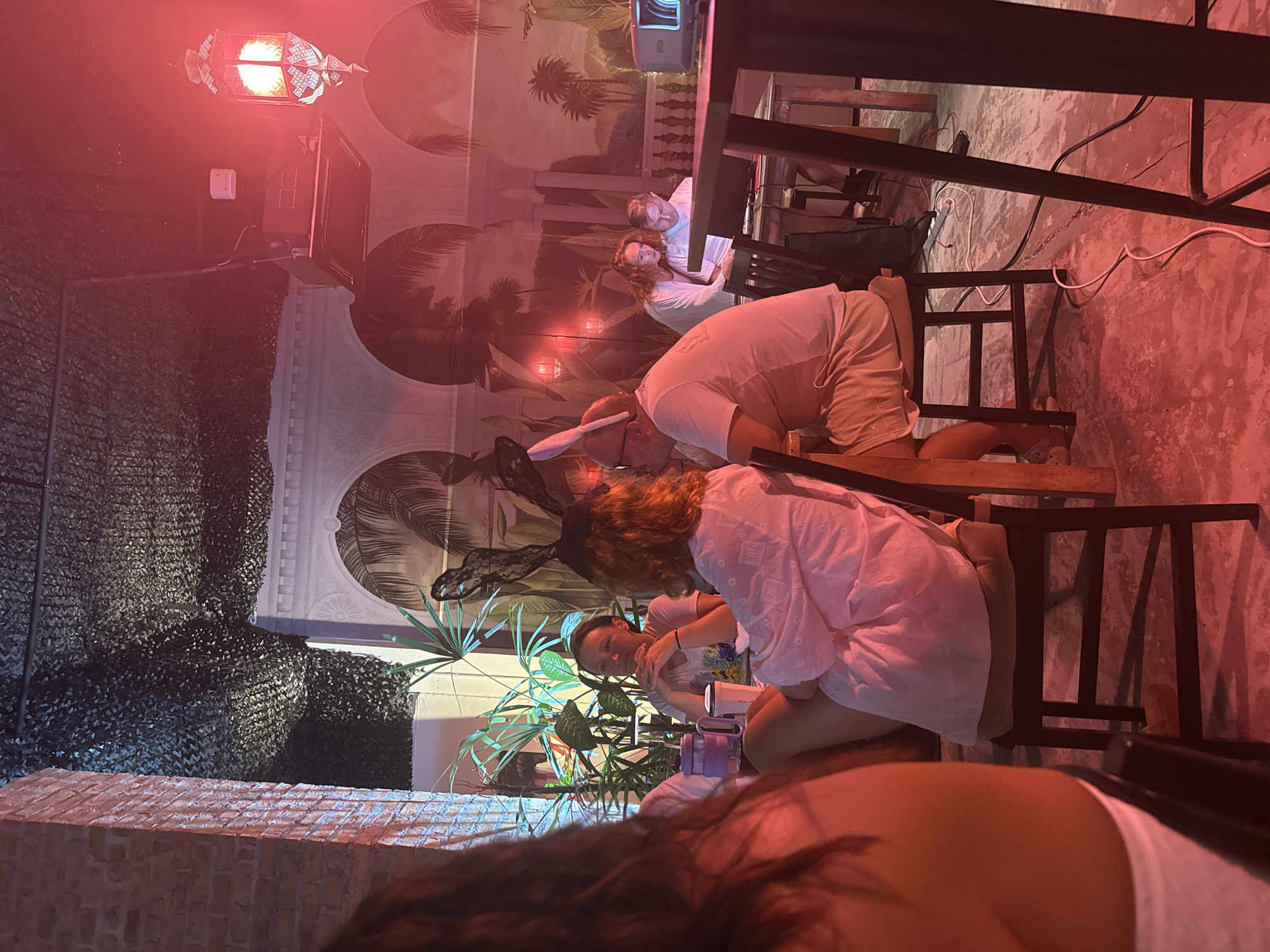The image shows a group of people gathered at what appears to be an outdoor seating area with a mural of palm trees on the wall. The lighting is predominantly red, possibly from a lamp or effect, casting a warm glow over the scene. Two individuals are seated at a table, one wearing what looks like bunny ears, suggesting a playful or themed gathering. Another person, with red hair, is leaning in towards the person with the bunny ears. In the background, two other individuals are visible, one wearing a white shirt. The setting includes tables and chairs, some with a metal frame. There's also a brick wall in the foreground and some greenery, possibly plants. The overall atmosphere seems relaxed and social, possibly an evening event due to the lighting. No text is clearly visible in the image.