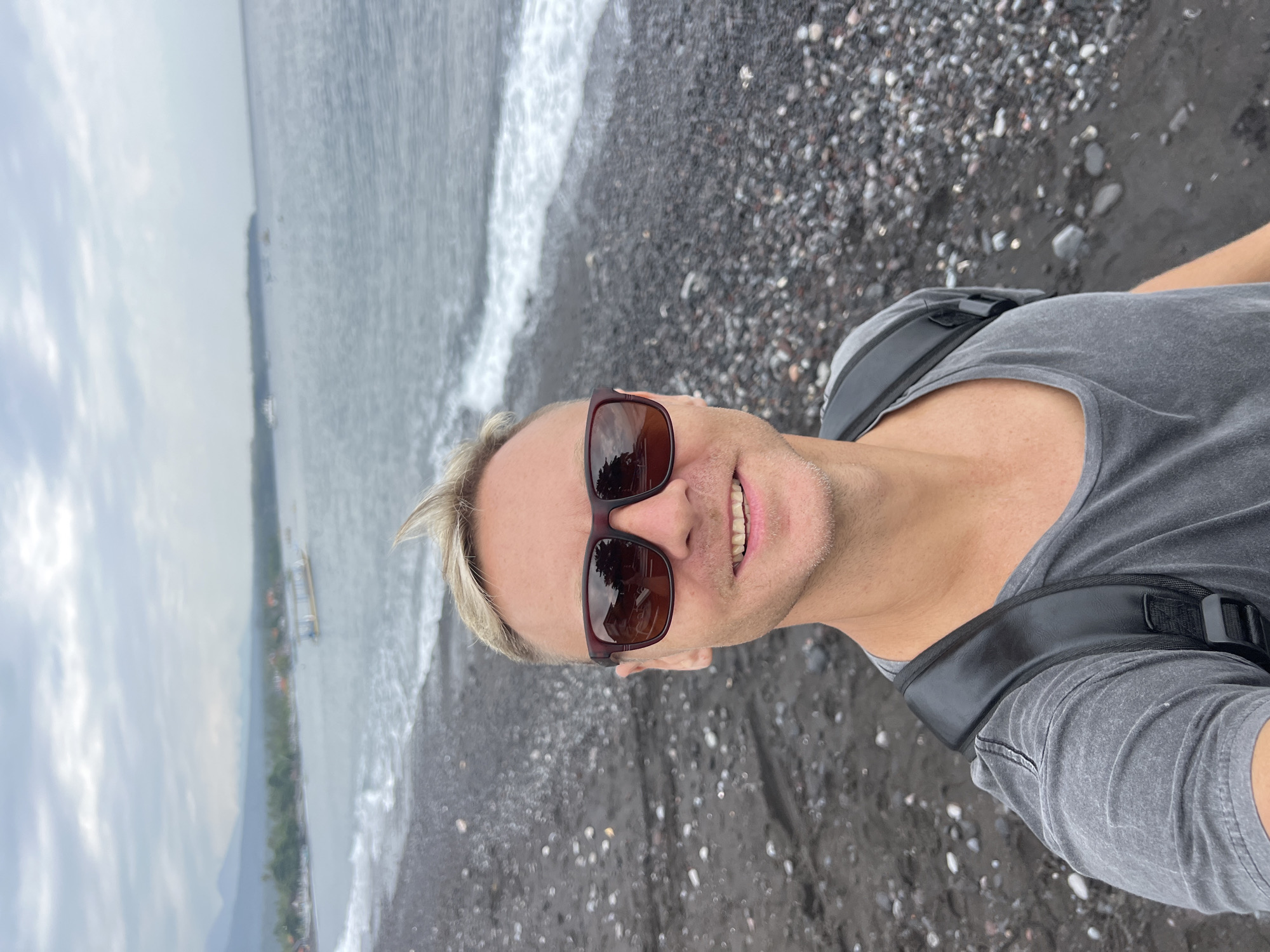 A medium shot captures a man with blond, spiky hair and sunglasses from the chest up, smiling and taking a selfie on a black pebble beach. He wears a grey t-shirt and a black backpack. The background shows the ocean with gentle waves, a coastline with lush green vegetation, and distant mountains under a cloudy sky. Several boats are visible on the water near the shore. This scene is located on the black sand beach of Abang, Indonesia.