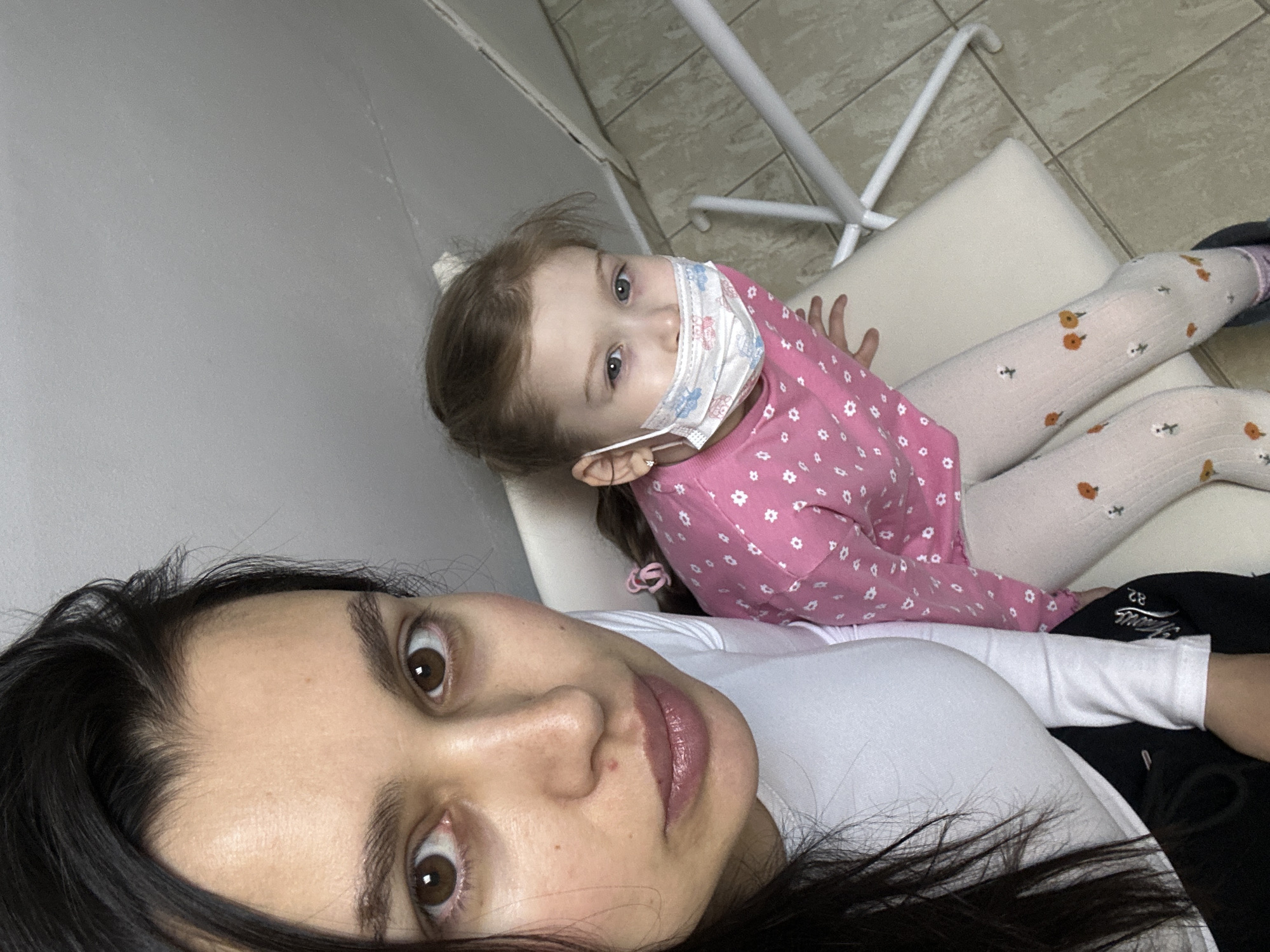 An indoor selfie features an adult woman and a young child. The woman, with dark hair and wide brown eyes, is in the foreground, looking directly at the camera. She wears a white long-sleeved top. Behind and slightly above her, a young girl with light brown hair and blue eyes looks calmly towards the camera. The girl is wearing a pink long-sleeved shirt with white floral patterns, white patterned tights, and a blue and white disposable face mask. She is seated or leaning on a simple white padded bench with a metal frame. The background consists of a plain light grey wall and a light-colored tiled floor. The sterile environment, along with the child wearing a mask, suggests a clinic or waiting room in Chaikovsky, Russia. On a dark fabric visible next to the adult's arm, the numbers "82" and partially legible text, possibly "Horeas," are visible.