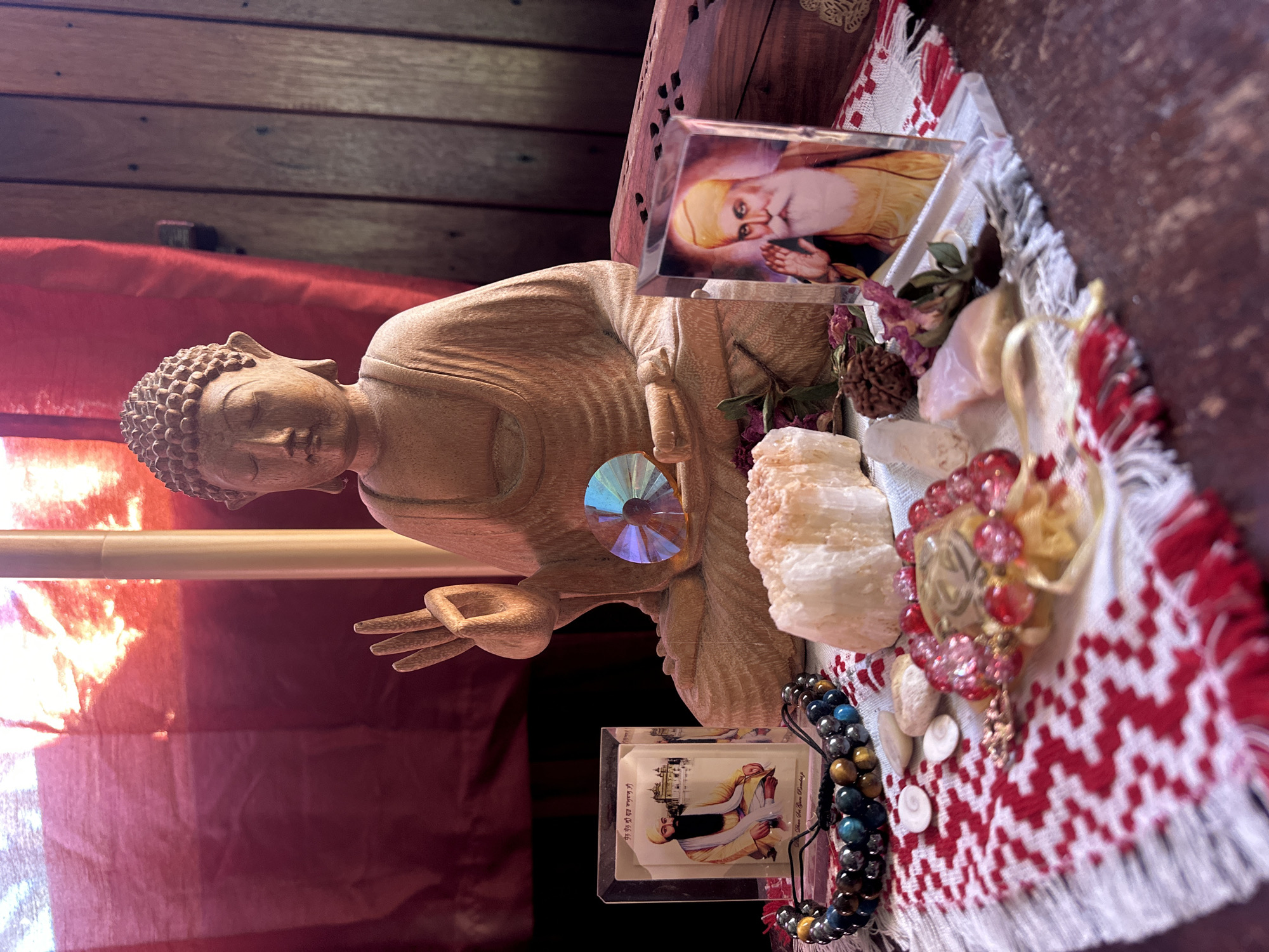The image depicts a close-up, eye-level, full shot of a wooden Buddha statue placed on a patterned cloth with red and white accents. To the left of the Buddha, a small framed picture shows a man in traditional attire seated in front of a building, and next to it, a beaded bracelet. In front of the Buddha, there are several crystals, some dried flowers, and a small glass ornament that refracts light. To the right of the Buddha, another framed picture shows a man with a long white beard and a yellow turban, with a wooden box behind him. The background features a red translucent fabric illuminated from behind, and a wooden wall on the right. The overall scene suggests a spiritual or meditative setting, possibly a home altar. The provided image does not contain any information about the city of Thora, Australia.