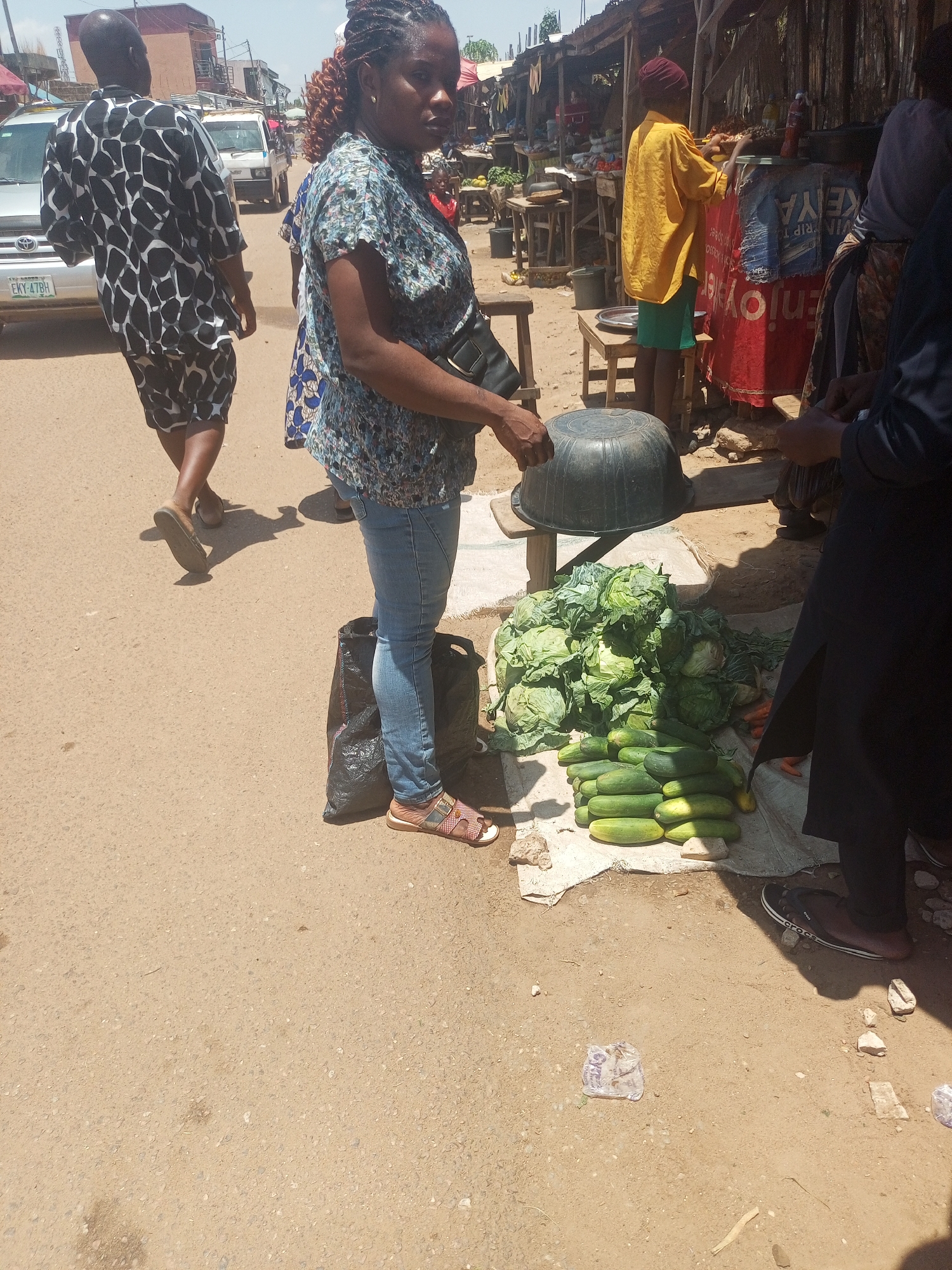 The image depicts a street scene in Sabon Tasha, Nigeria. A woman in a floral shirt and jeans stands next to a display of cabbages and cucumbers laid out on a white cloth on the ground. A black plastic bowl sits on a stool nearby. Other individuals are present: a man in a patterned shirt walking away from the camera, a person in a yellow shirt behind a red stand, and another individual partially visible on the right. A car with license plate "EKY-478BH" is visible in the background. The environment suggests an outdoor market with stalls lining the sides of the street.