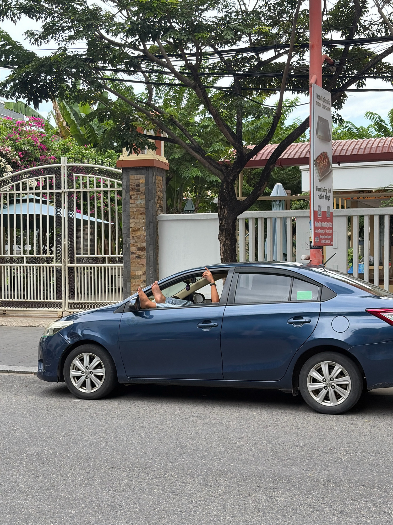 A dark blue sedan is parked on the side of a street in Đà Nẵng, Vietnam. The driver's side window is open, and a person's bare feet are propped up on the windowsill, with their toes visible. The car is parked in front of a white metal gate with decorative scrollwork, behind which are lush green trees and flowering bushes with pink and white blooms. A stone pillar with a decorative brown cap stands to the right of the gate. A red pole with a sign advertising pizza is visible in the background, mounted on the sidewalk. The street is paved with asphalt, and the sky is overcast.