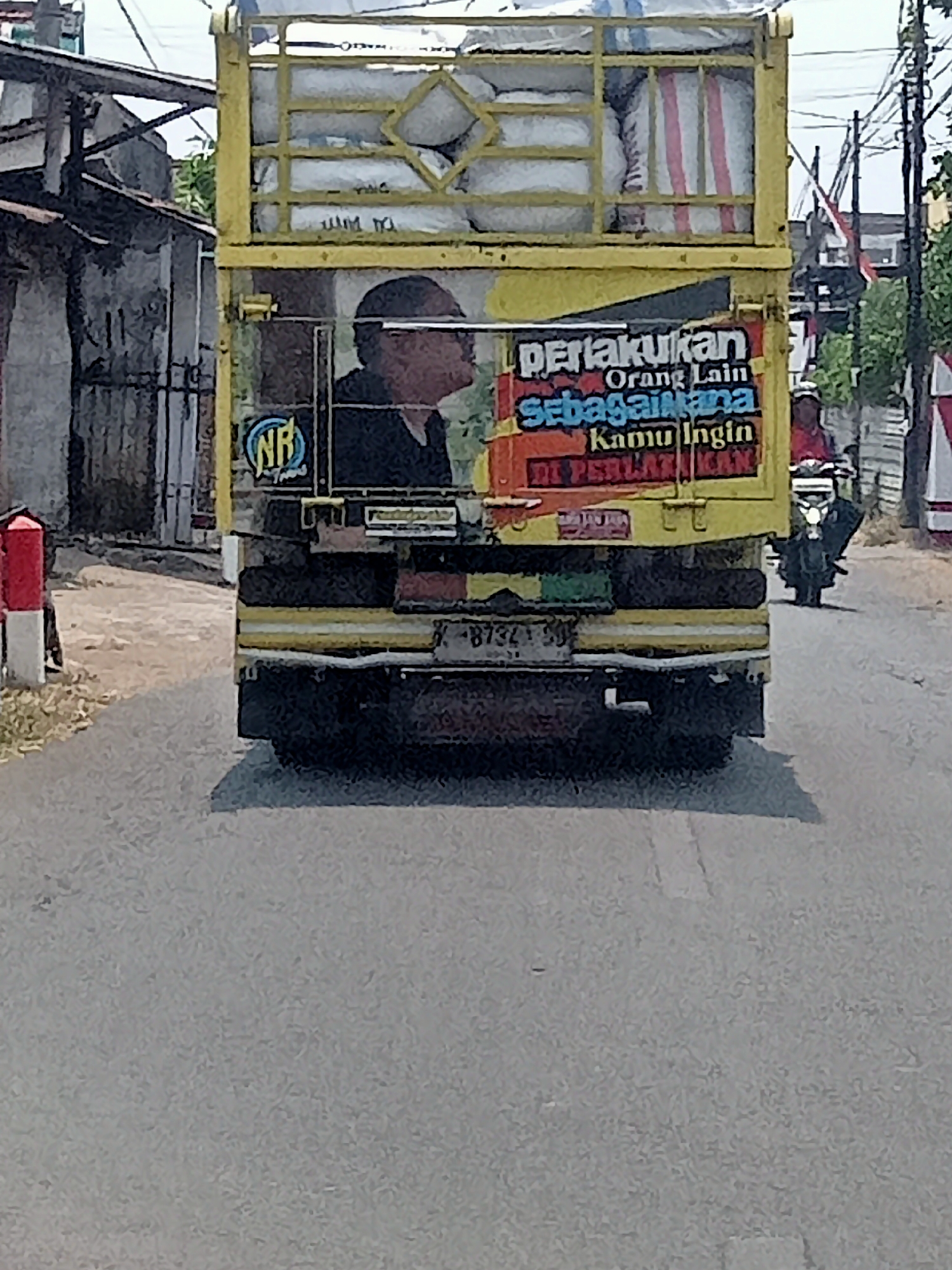 A rear view of a yellow truck is captured on a street in Simo, Indonesia. The truck is laden with white sacks, some of which are wrapped in plastic. A yellow frame structure is visible above the truck bed, and diagonal yellow bars are present on the rear of the truck. A decal on the truck displays text in Indonesian, reading "pertakukkan Orang Lain sebagaimana Kamu Ingin Di Perlakukan." A circular logo with the letters "NR" is also affixed to the truck. To the left of the truck, a red and white striped pole stands beside a building with a corrugated metal roof and a weathered facade. In the background, other buildings and trees are visible. A motorcycle with its headlights on is to the right of the truck, and a person is seated on it. The road surface is asphalt, with some scattered debris. The overall scene appears to be in daylight.