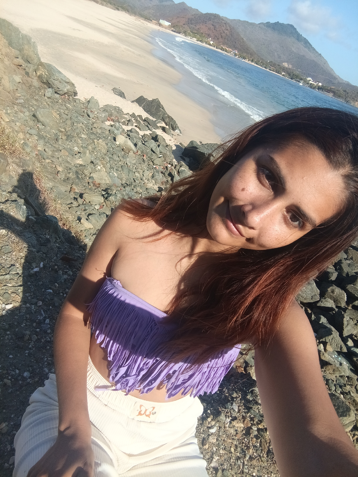 A young woman with brown hair and fair skin with freckles takes a selfie on a rocky beach. She is wearing a purple fringed bikini top and white pants. Behind her, a sandy beach meets the blue ocean, with waves gently lapping the shore. In the distance, a range of green mountains rises under a clear blue sky. The sun casts a shadow of the woman on the rocks.