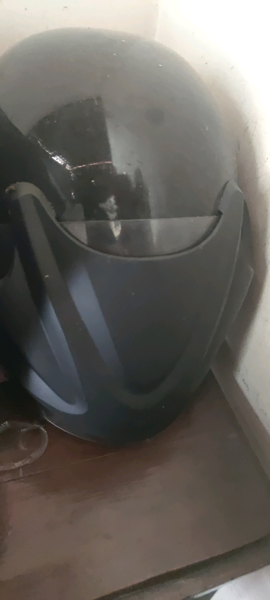 A black motorcycle helmet is pictured in the city of Gambarsari, Indonesia. The helmet is made of a glossy black material and has a clear visor. It is positioned on a wooden surface, likely a table or shelf, which appears to be dark brown and reflects some light. The background consists of a wall, which is a light color and shows some texture and faint markings. To the left of the helmet, a portion of a circular, metallic object is visible, possibly part of a stand or another item. The lighting in the scene is somewhat diffused, creating shadows and highlights on the helmet's surface. The overall environment suggests an indoor setting, possibly a storage area or a room in a house.