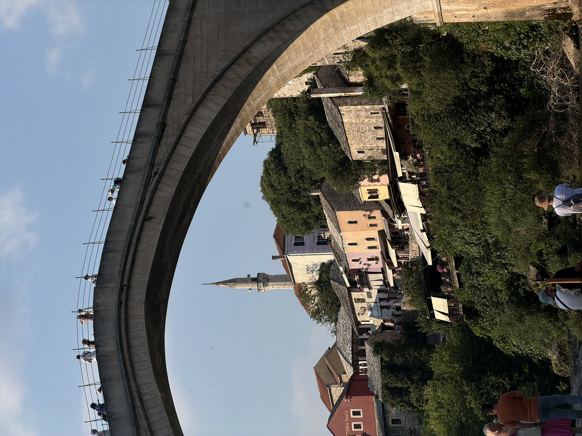 The image shows a view of the Stari Most (Old Bridge) in Mostar, Bosnia and Herzegovina, from below and to the side. The stone arch of the bridge dominates the upper portion of the frame. On the bridge, several people are visible, standing and looking out. Below the bridge, a cluster of historic buildings, characterized by stone and stucco facades and red-tiled roofs, cascades down a hillside. A minaret of a mosque rises above the buildings. Lush green trees and vegetation cover the slopes. In the foreground, near the bottom of the image, several people are gathered by what appears to be a river or body of water. One person is wearing an orange t-shirt and jeans, another a pink dress, and another a hat. The sky is a clear blue with a few scattered clouds.
