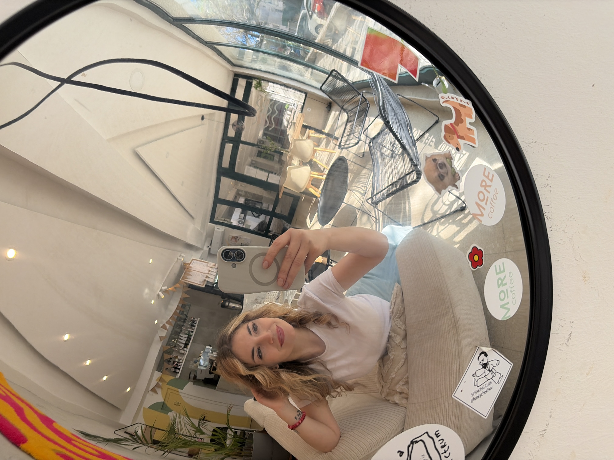 The image shows a reflection in a round mirror with a black frame. A young woman is holding a smartphone and taking a selfie. The mirror is decorated with stickers, including one that says "MORE coffee," a flower, and a dog. The reflection shows a cafe interior, likely in the city of Budva, Montenegro. The cafe has a glass roof, light-colored walls, and various tables and chairs. The woman is sitting on a light-colored cushioned chair. The reflection shows decorative lights, plants and menu boards.