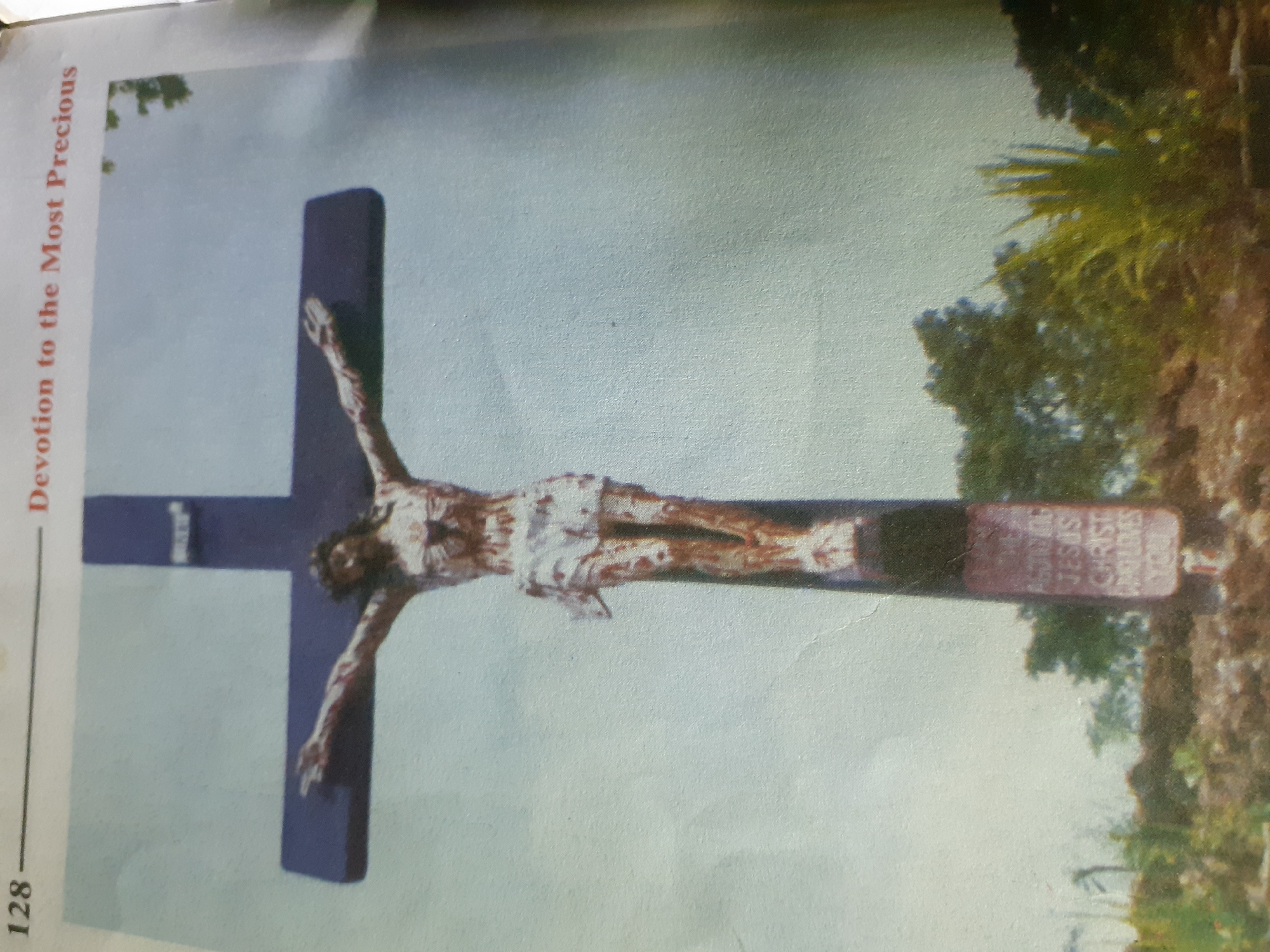A full-body statue of Jesus Christ is depicted on a cross, positioned outdoors. The statue is made of a brown material, with white cloth wrapped around Jesus's waist. Jesus's arms are extended horizontally, and his head is bowed. The cross is dark blue and stands on a brown stone base, which is inscribed with white lettering that reads: "AMAZING JESUS CHRIST WHO LOVES YOU." The background is a hazy, light blue sky with some green trees visible in the lower half of the image. The image also shows a page number "128" and the text "Devotion to the Most Precious" at the top. It is not possible to determine the exact location in Jalingo, Nigeria from the image.