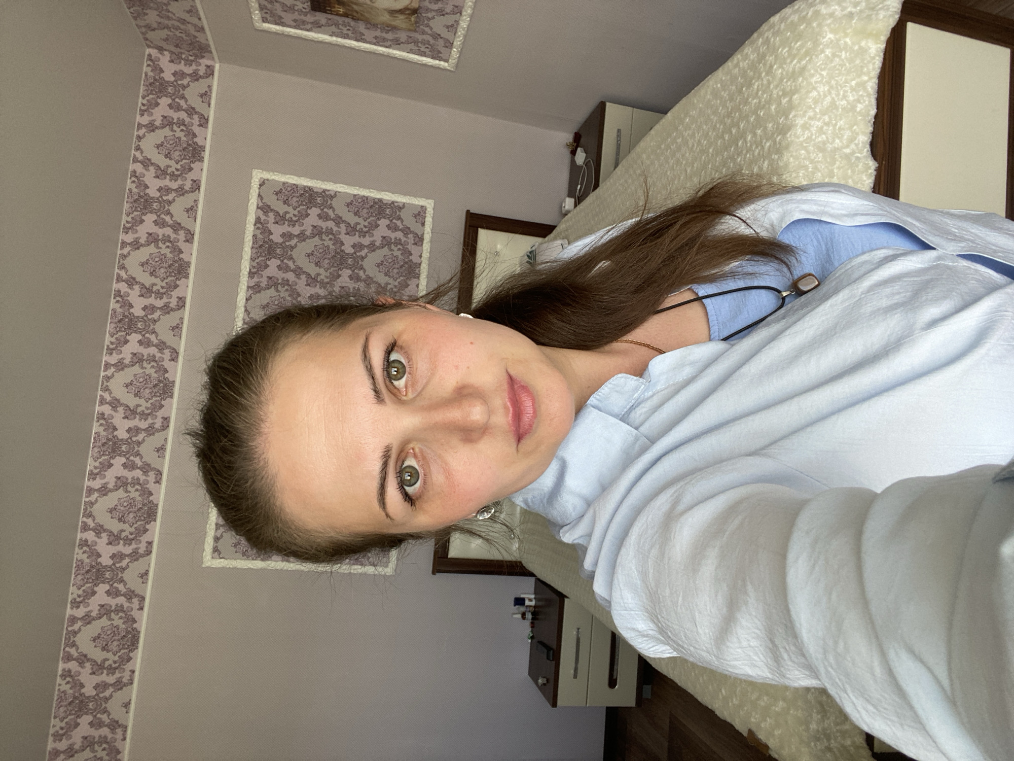 A woman with light brown hair tied back in a ponytail takes a selfie in what appears to be a bedroom. She is wearing a light blue collared shirt open over a blue tank top and a pendant necklace. Her left hand is extended, holding the camera. The background shows a bed with a cream-colored textured comforter, two nightstands with lamps and various items on them, and wallpaper with a floral pattern. The image does not provide enough information to determine a specific location within Марковский, Russia.