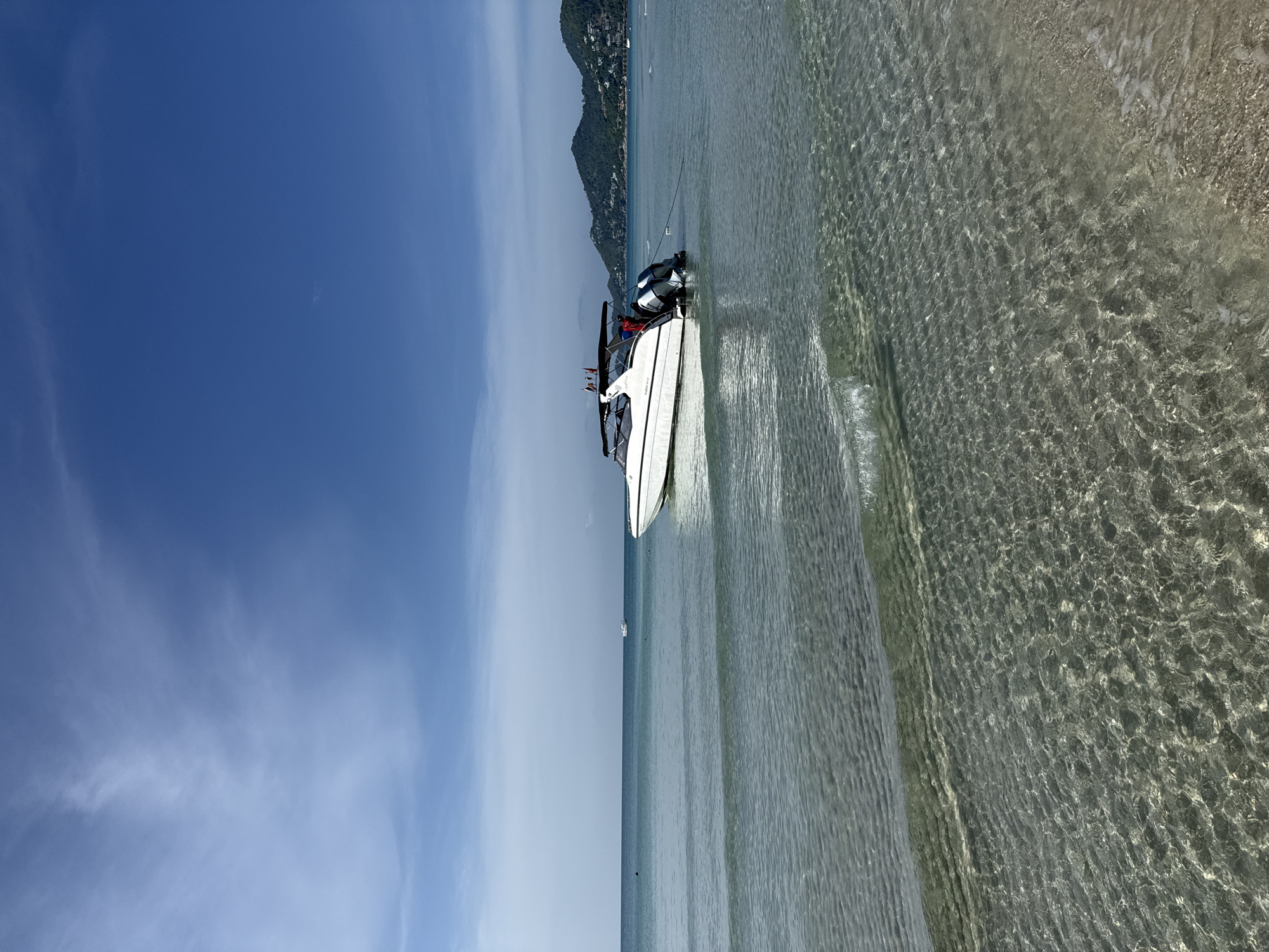 This is a photograph taken at a beach in the Ko Samui District of Thailand. The image captures a serene coastal scene with a clear blue sky and calm, turquoise waters. In the foreground, the shallow water gently laps against the sandy shore, with sunlight creating a shimmering effect on the seabed. A sleek white speed boat is anchored in the mid-ground, its twin outboard motors visible. Two individuals, wearing red and black tops, are standing on the boat, seemingly engaged in an activity. In the distance, a chain of lush green hills, characteristic of Ko Samui's topography, forms the backdrop, with some structures visible on their slopes. The overall atmosphere is one of tranquility and a typical tropical island setting.