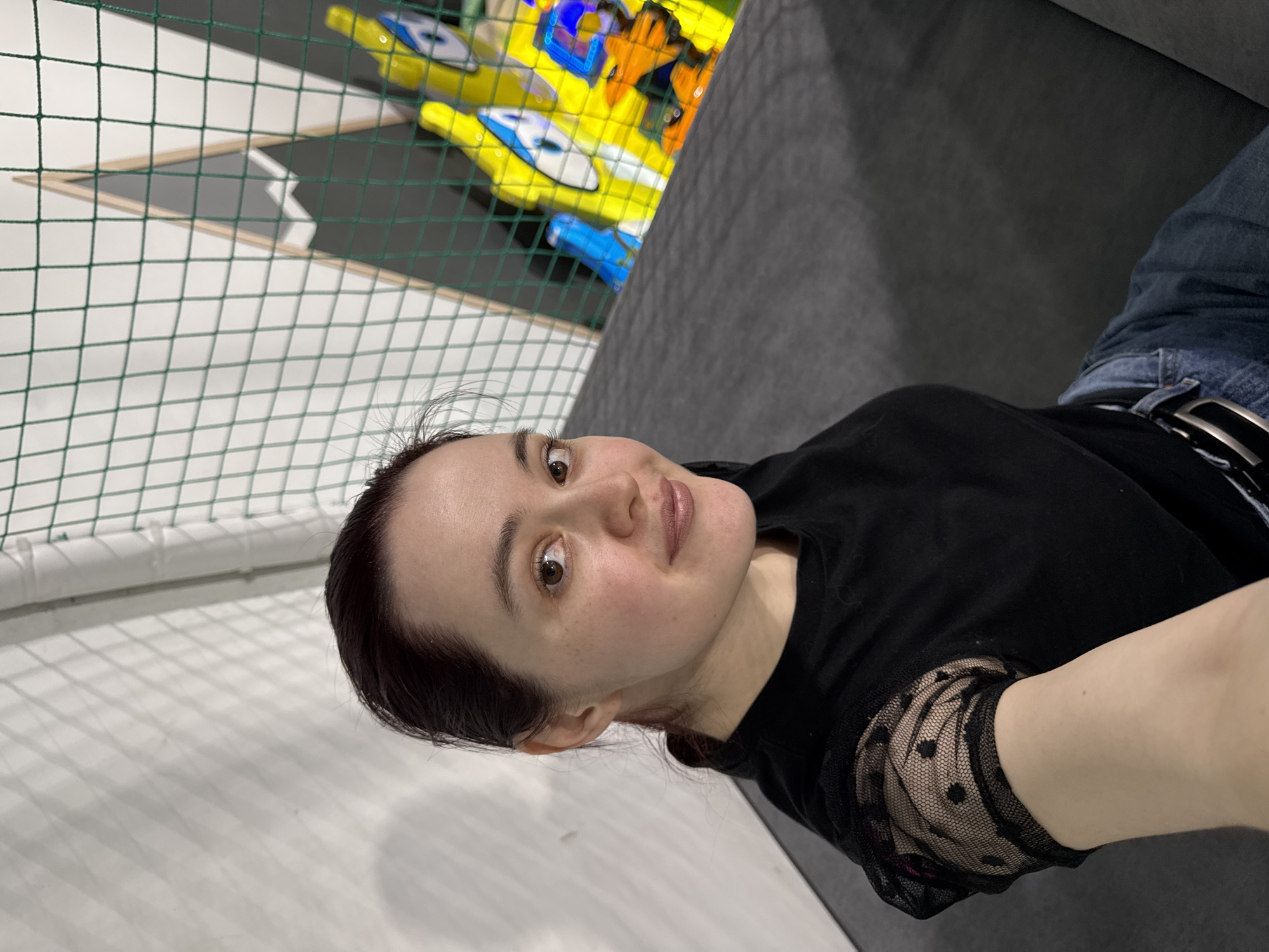 A woman is captured in a selfie taken indoors, looking directly at the camera with a calm, slightly smiling expression. The image is rotated 90 degrees clockwise. She has dark hair and brown eyes, with a light complexion and visible freckles. She is wearing a black short-sleeved top with distinctive sheer, polka-dotted sleeves, blue jeans, and a black belt with a silver buckle.
The setting appears to be a children's indoor play area in Izhevsk, Russia. A green safety net is prominent in the background, separating the woman from colorful play structures. Behind the net, several brightly colored plastic elements are visible in shades of yellow, blue, and orange, resembling small boats or vehicles often found in soft play zones or ball pits. A dark gray padded surface is also present in the background, likely part of the play area's floor or equipment. To the woman's left (bottom in the current orientation), a white padded wall or floor can be seen, partially covered by the safety net. The environment suggests artificial lighting. No visible text is present in the image.