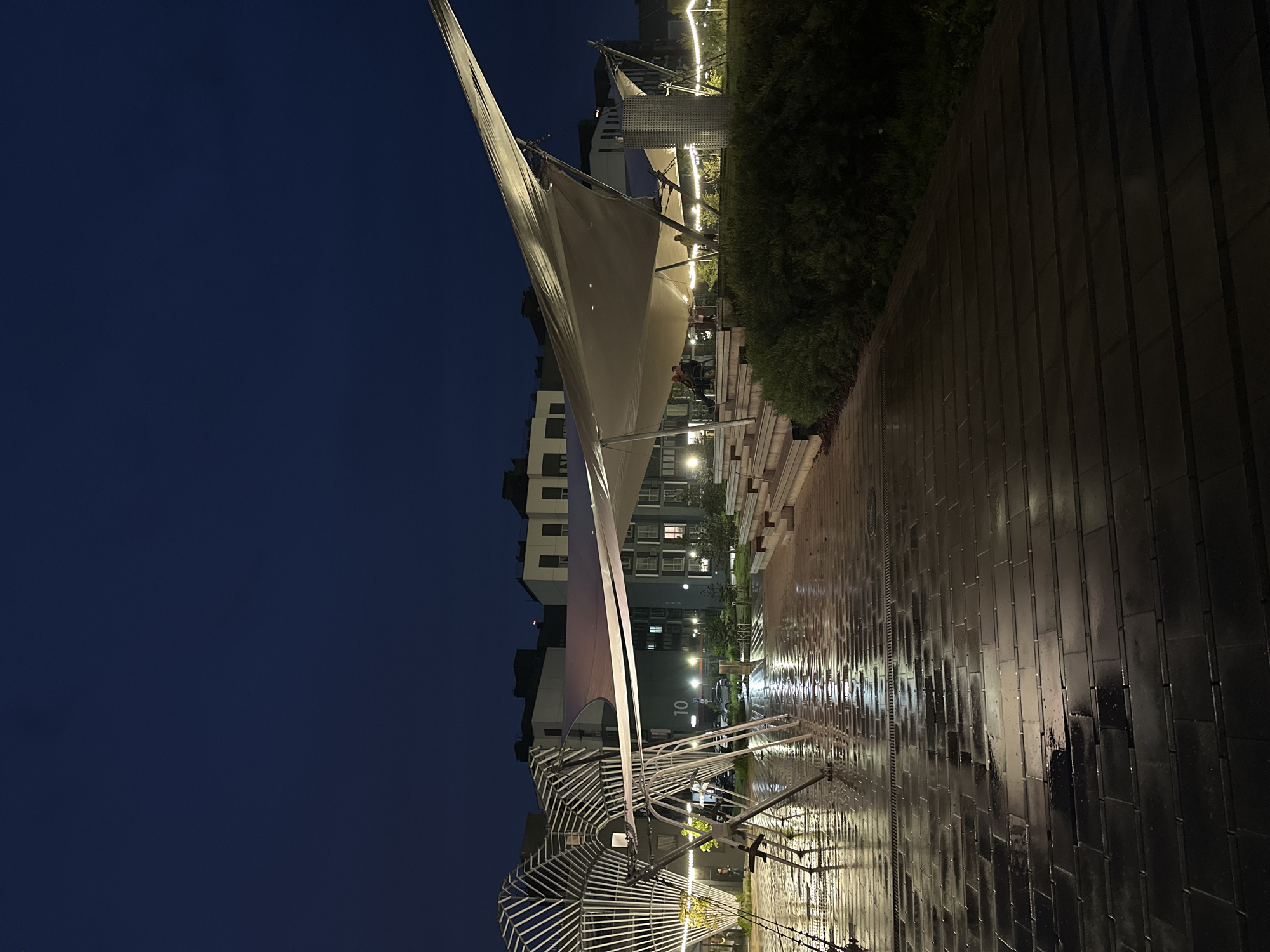 The image captures a nighttime scene in Бараўлянскі сельскі Савет, Belarus, likely taken after a recent rain, as evidenced by the wet pavement. In the foreground, the wet walkway reflects the ambient light, creating a shimmering effect. A large, white, tensile structure dominates the scene, supported by a network of white metal poles and cables, possibly a canopy or shade structure. In the background, multi-story apartment buildings are visible, their windows emitting light. The overall lighting suggests a well-lit urban environment at dusk or early evening. Some greenery in the middle ground hints at a park or landscaped area. No individuals are clearly visible, but the presence of seating under the tensile structure suggests it is a public space intended for gathering.
