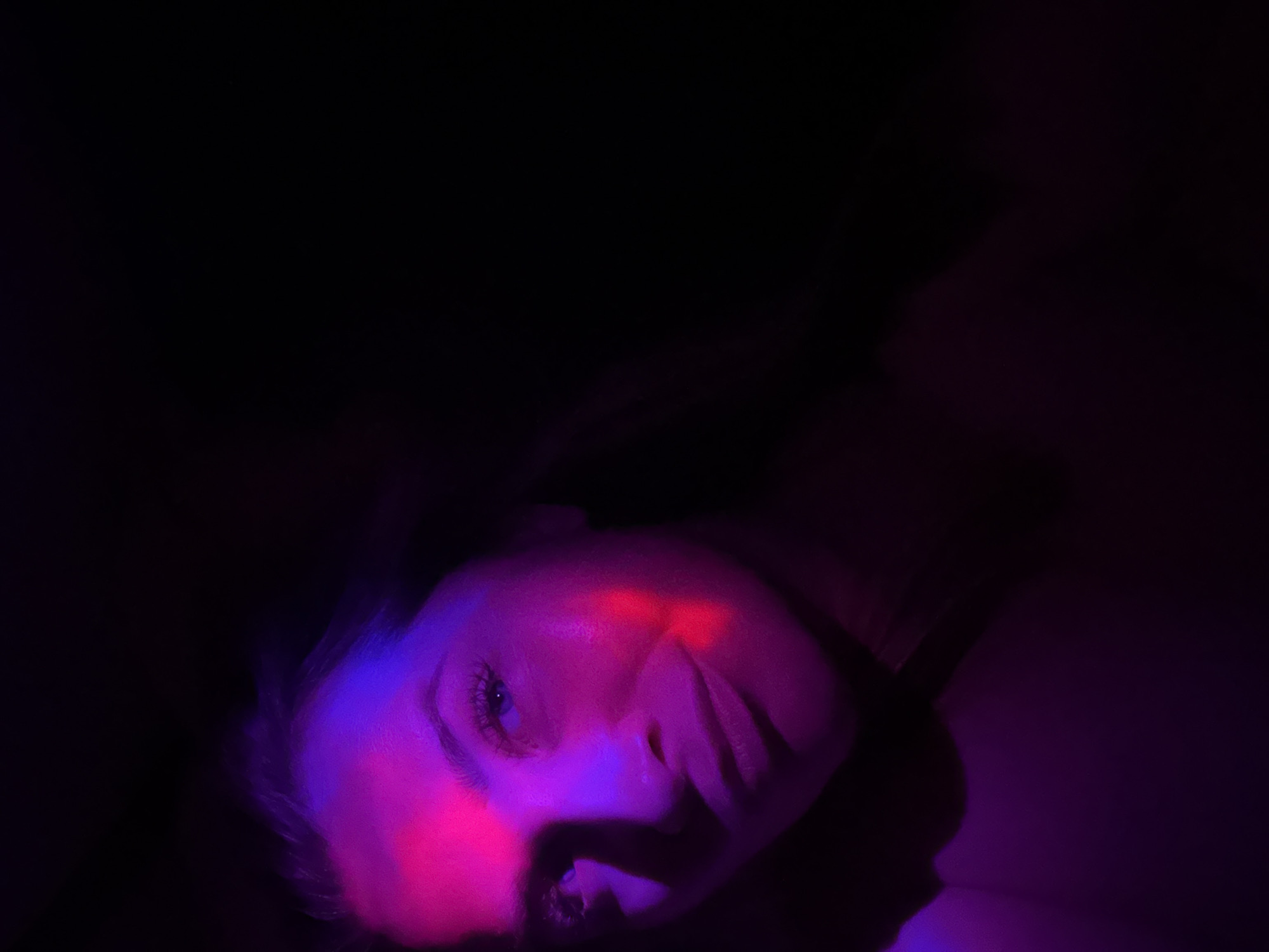 The image is a close-up, low-angle portrait of a young woman's face, illuminated by dramatic purple and pink lighting against a dark background. The woman's eyes are open and directed towards the camera. A streak of red light crosses her cheekbone. The background is predominantly black, with some ambient purple light visible on the left and bottom edges of the frame. The image is underexposed, with significant shadow obscuring parts of the woman's face and hair. There are no discernible features of the city of Veliky Novgorod, Russia, or any other external environment.