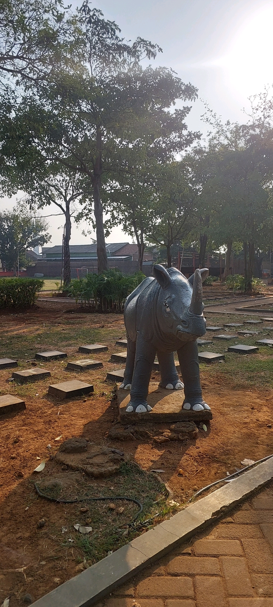 This is an outdoor, full-shot photograph taken at eye-level. The scene depicts a concrete statue of a rhinoceros in a park-like setting. The rhino statue is dark gray, with white accents on its feet and horn. It is positioned on a raised concrete platform in the foreground, surrounded by reddish-brown dirt and patches of grass. Several square, flat concrete pavers are laid out in a scattered pattern on the ground around the statue.
In the background, there are several large trees with full, green canopies, casting shadows on the ground. Beyond the trees, there are buildings with visible roofs, suggesting an urban or suburban environment. The sky is bright and hazy, with the sun appearing to be high in the sky, indicated by the bright light and lens flare in the upper right portion of the image. The overall impression is of a public park with a decorative statue. The scene is consistent with the location of Taman Mpu Tantular in Semarang, Indonesia, which features similar rhinoceros statues and park elements.