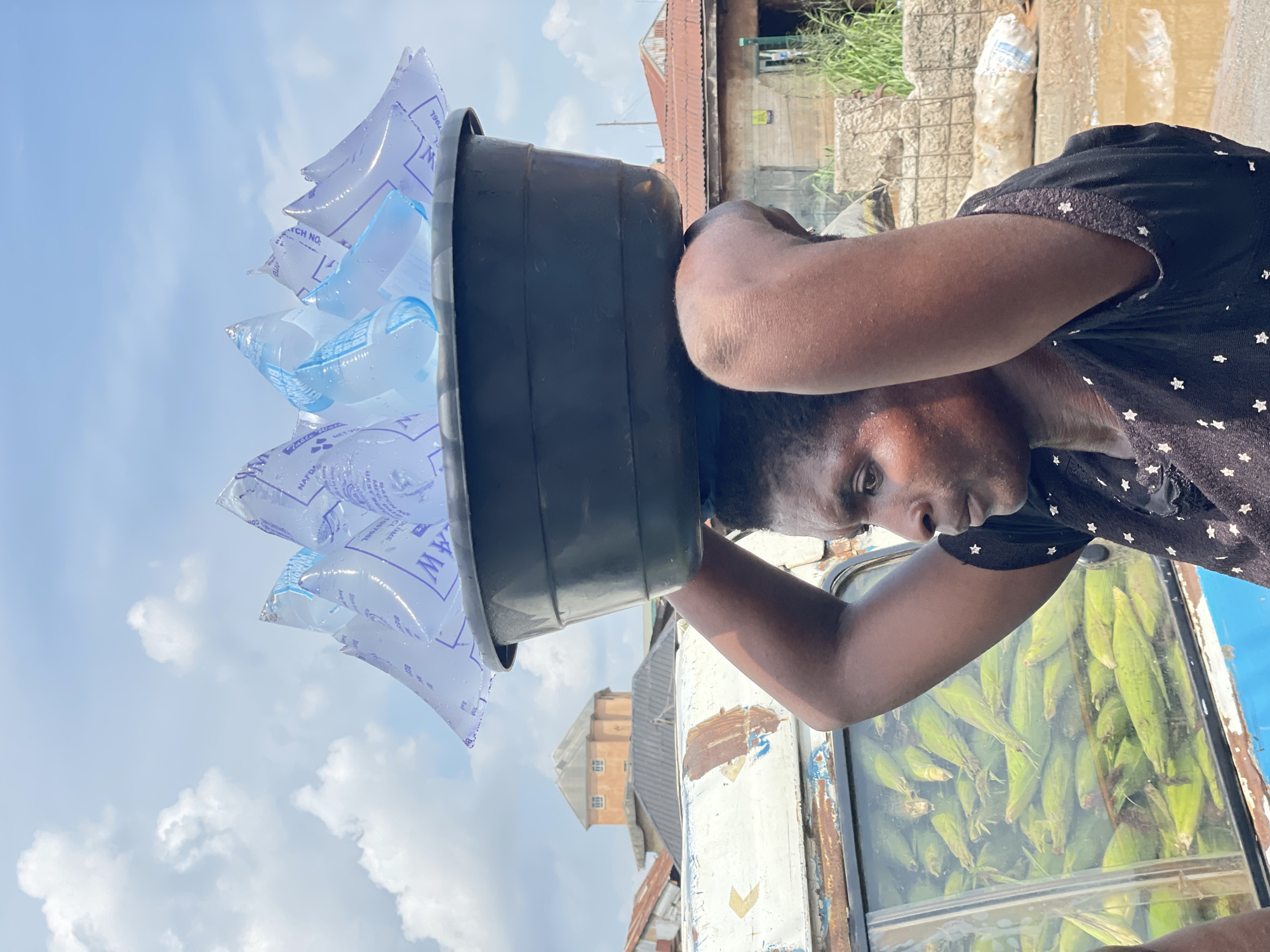 A low-angle shot captures a person, likely an adult with dark skin and short dark hair, carrying a large black plastic basin filled with numerous transparent water sachets on their head. The person is positioned on the right side of the frame, looking towards the left with a neutral, focused expression. Their left arm is visible, bent at the elbow.
The basin is overflowing with the individual sachets of drinking water, which glisten in the bright light. Visible text on some of the sachets includes "NAFDAC", "BATCH NO.", "VOLUME", and partial brand indicators such as "MW" or "AMW," along with numbers like "1964" or "1981."
In the background, immediately behind the person, is what appears to be a market stall or cart, with a glass panel showcasing a dense display of fresh green corn cobs. Beyond this, the scene unfolds into an outdoor urban or semi-urban environment under a clear blue sky dotted with scattered white clouds, indicating a bright, sunny day. Various buildings with corrugated iron roofs and stone or concrete walls are visible, along with some green vegetation. A distinctive multi-story building, possibly a tower, stands out in the distant background. The image depicts a common street vending activity.