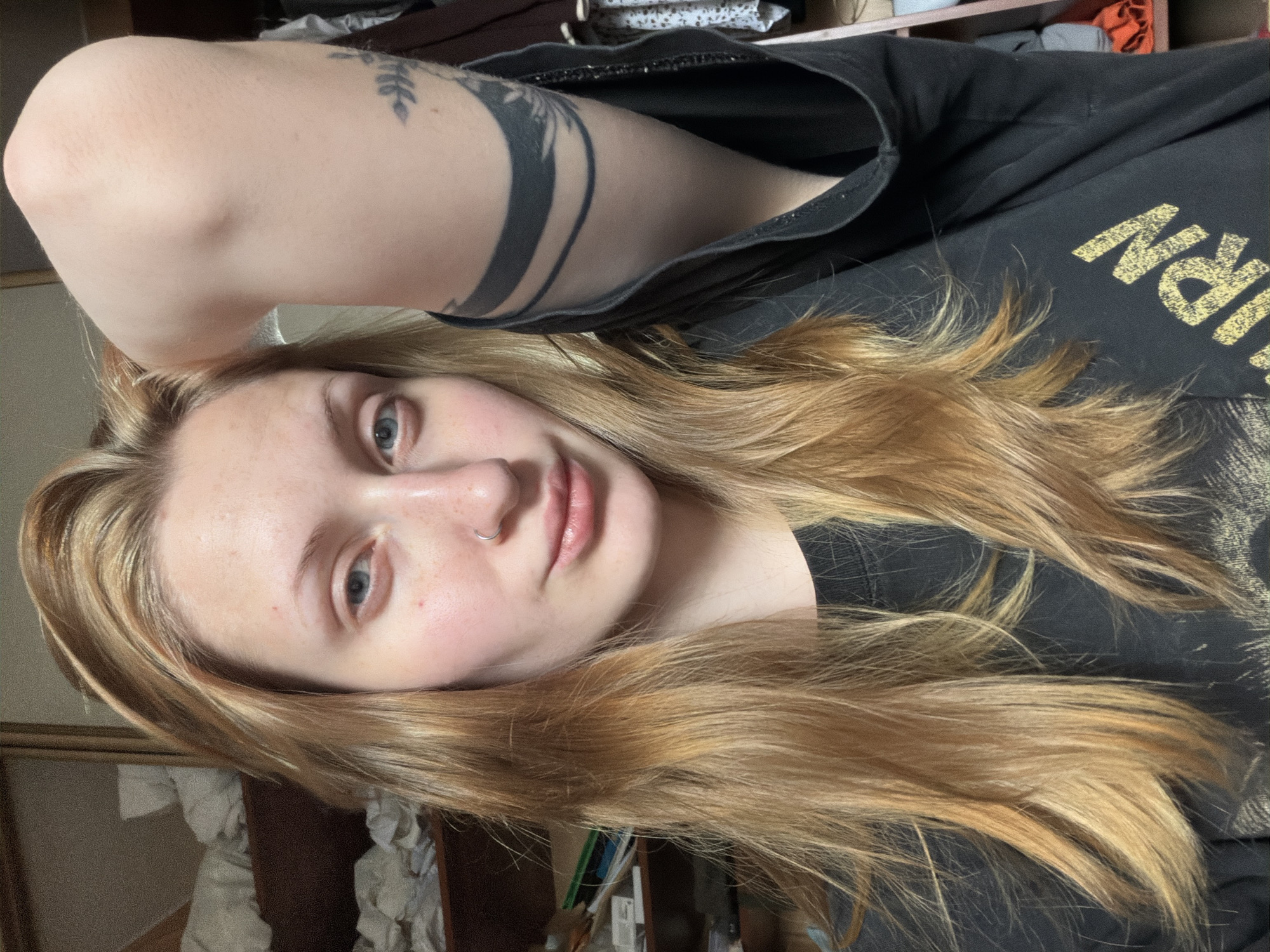 A young woman with blonde hair and blue eyes is captured in a selfie. She is wearing a black t-shirt and has a nose ring. A tattoo is visible on her right arm. The background appears to be an indoor setting, possibly a bedroom, with shelves filled with various items. The overall lighting suggests an indoor environment. No specific location in Ladozhskoe Ozero, Russia can be identified from the image.