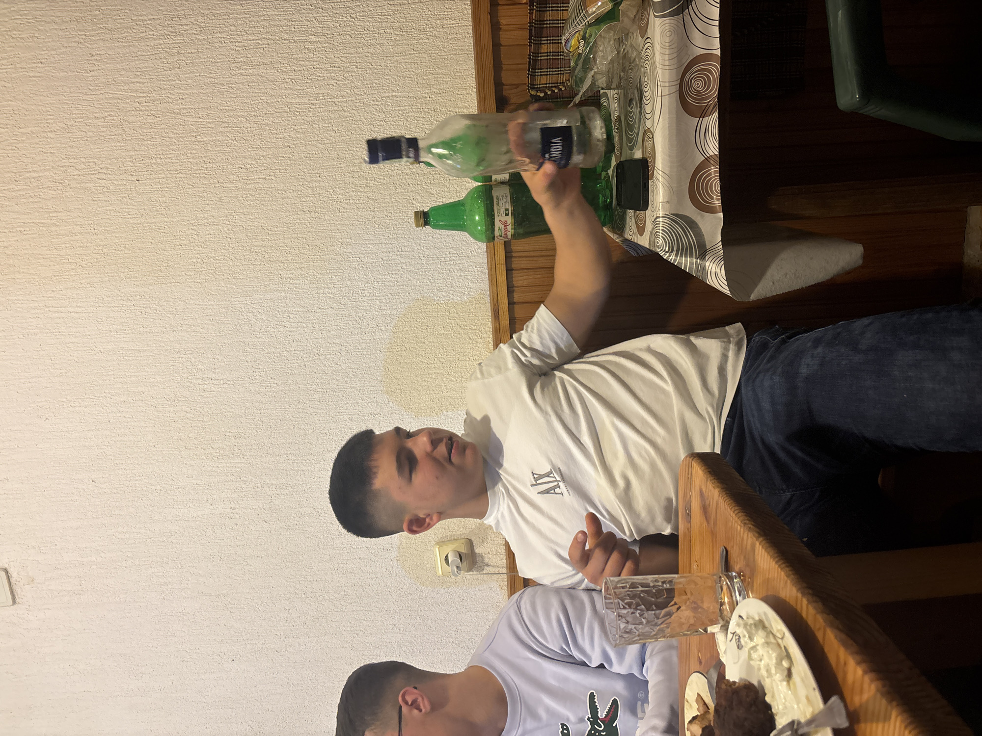 The image shows two men at a table. One man is holding a bottle of vodka. There are also bottles of "Mastika" at the table. The location is likely a home in Zlatograd, Bulgaria. Food and drinks are present on the table. The setting appears to be casual and social.