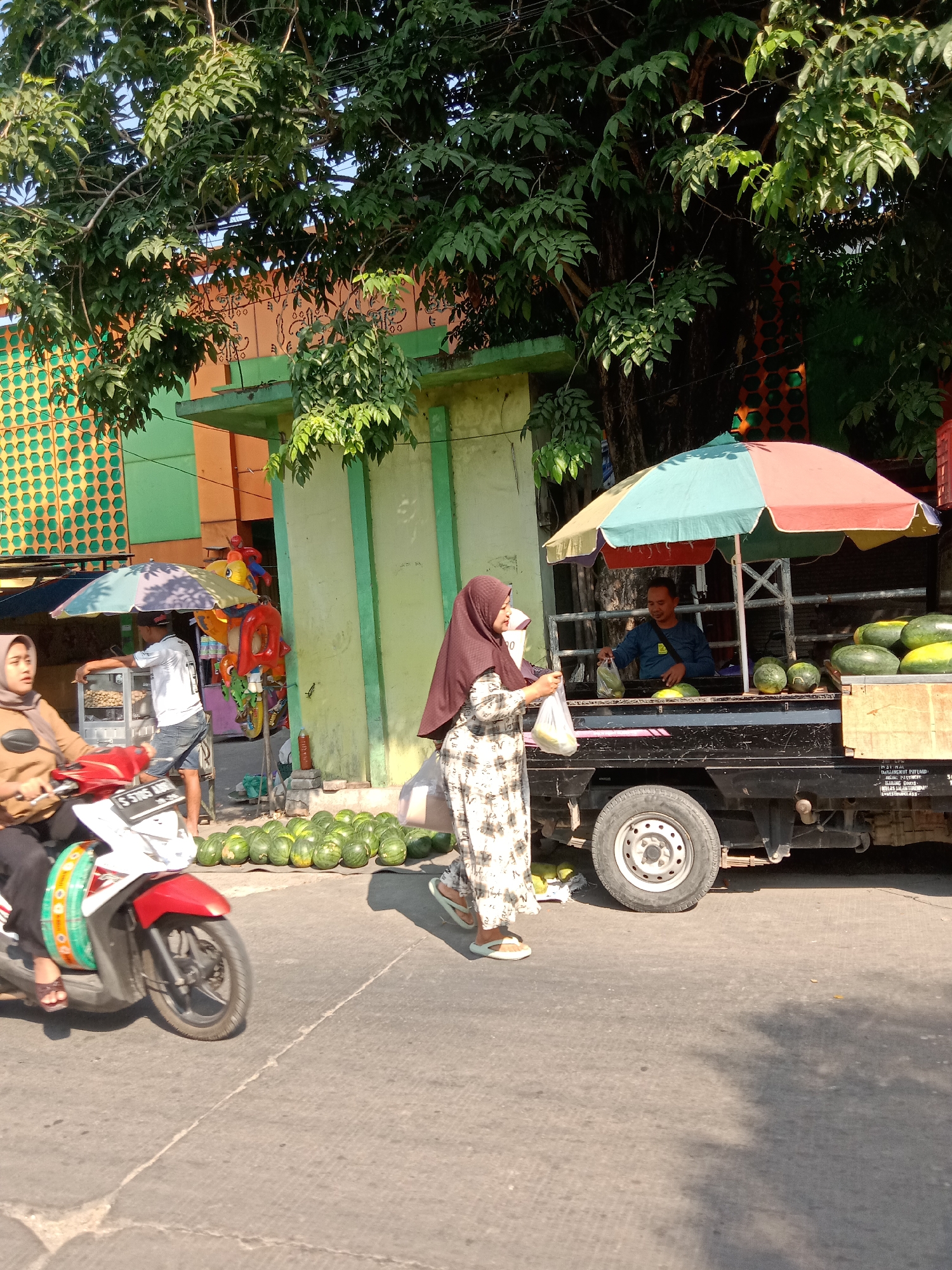 In the city of Sugihwaras, Indonesia, this image captures a street scene with a watermelon vendor operating from the back of a truck. A man, presumably the vendor, is visible behind a colorful umbrella, interacting with a customer. He is wearing a blue shirt and appears to be handing a plastic bag to the customer, who is a woman dressed in a patterned dress and a headscarf. In the foreground, a woman wearing a headscarf is riding a red and white motorcycle. The truck is loaded with watermelons, and more watermelons are displayed on the ground in front of it. The environment suggests a sunny day, with shadows cast on the road, and lush green trees overhead. Behind the vendor's truck, a building with green and orange sections is visible, along with decorative elements. The overall scene depicts a typical day of commerce on a street in Indonesia.