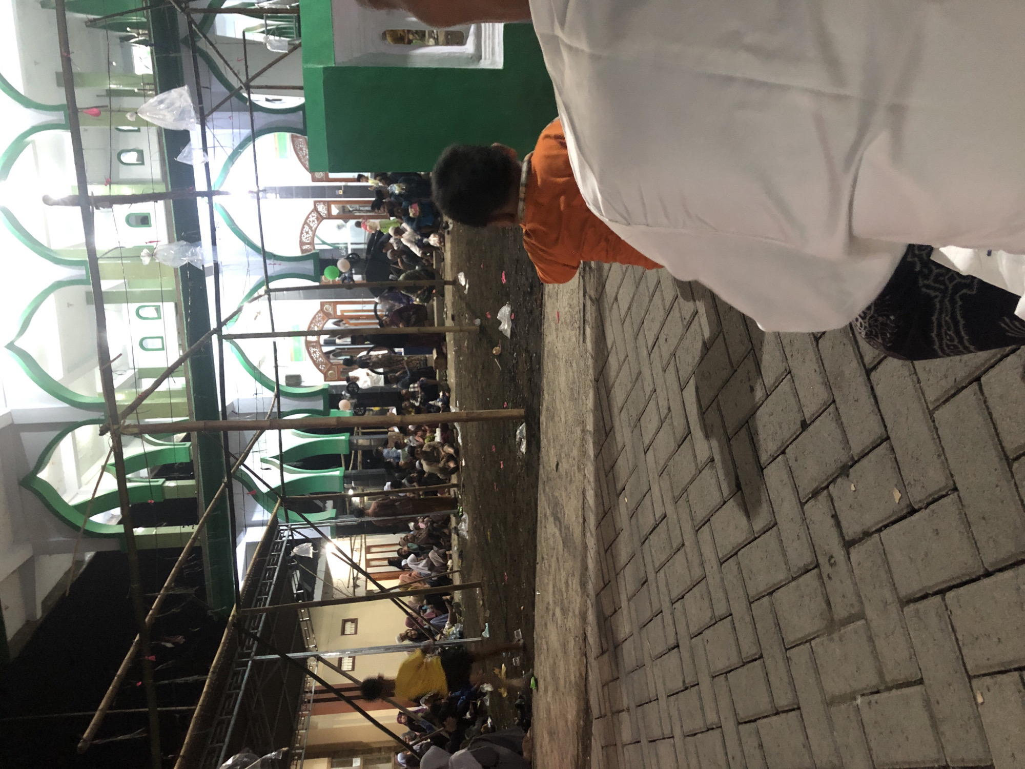In Tenggilis Rejo, Indonesia, this image captures an outdoor gathering at night, likely near a mosque given the architectural elements. A crowd of people, dressed in traditional attire, are seated on the ground amidst temporary wooden structures and scaffolding. Some individuals are standing, and a few children are visible. In the foreground, a child in an orange shirt is facing away from the camera, while a person in a white garment is also partially visible. The ground is a mix of paved stone and dirt. The scene suggests a communal event or religious observance taking place after dark.