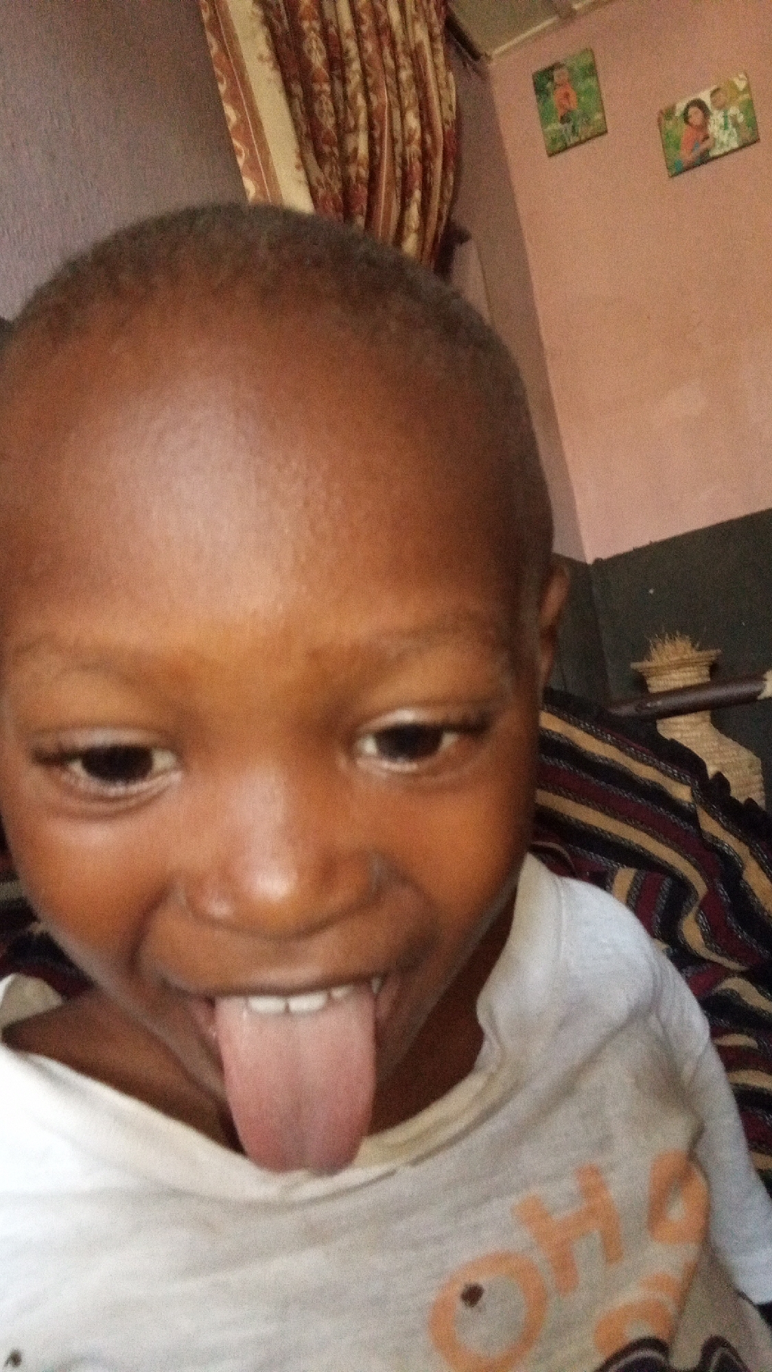 A close-up, slightly blurry image shows a young child, likely a toddler or preschooler, looking downward with a playful expression, their tongue sticking out. The child has dark skin and closely cropped dark hair. They are wearing a light-colored t-shirt, possibly white or off-white, with partially visible orange text that appears to spell "OH" on the front.
The background reveals an indoor setting. To the upper left, patterned curtains with vertical stripes in shades of red, brown, and cream are visible. A light pinkish-brown wall occupies the upper right side of the frame, where two small, framed pictures or canvases depicting people hang. Behind the child's right shoulder, part of a patterned fabric, possibly a blanket or upholstery, with stripes in black, maroon, and beige, can be seen. The lighting suggests an indoor environment, but the time of day is not discernible. The overall mood is playful and candid.