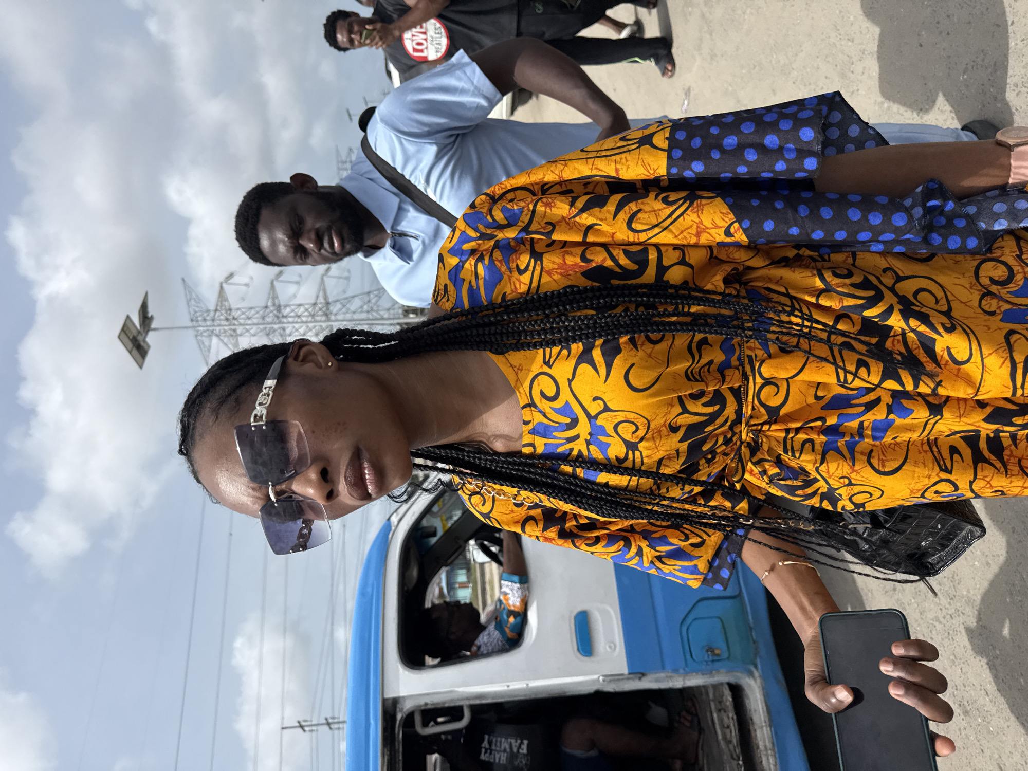 A woman with braided hair, wearing sunglasses and a yellow and blue patterned dress, stands near a blue and white bus, holding a black smartphone in her right hand. Behind her is a man in a light blue shirt and a black strap across his chest. In the background, another man is partially visible, wearing a dark shirt with a red and white circular design. The scene takes place outdoors, evidenced by the presence of overhead power lines and a partly cloudy sky. The location is identifiable as Lekki, Nigeria based on the recognizable style of the bus and the urban environment.