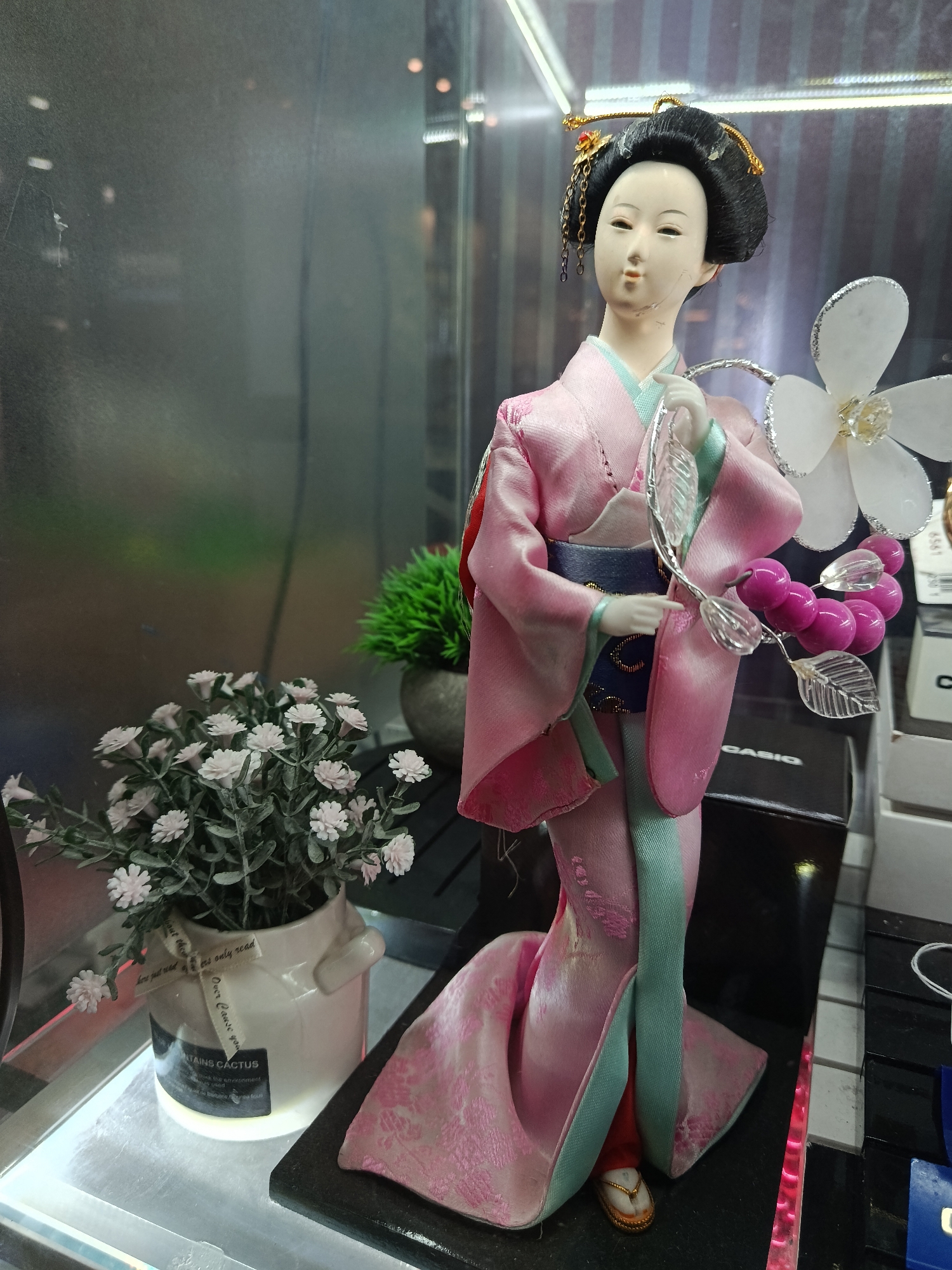 A geisha doll in a pink kimono stands in what appears to be a glass display case, surrounded by various items, including a potted plant with white flowers, a green bush in a grey pot, and a Casio watch box. The doll holds a decorative branch with pink berries and a white flower. Text is visible on the flower pot, and the background includes reflections of light sources, suggesting an indoor environment. The location in the city of Moscow, Russia is unknown.