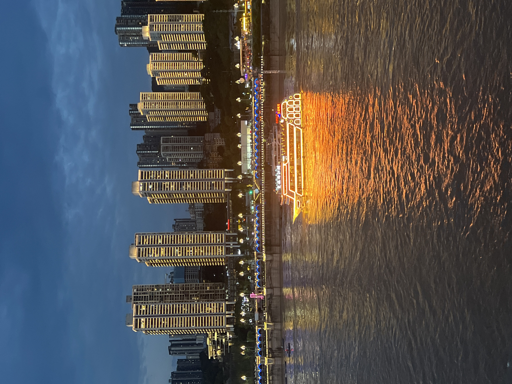 The image depicts a nighttime view of a waterfront cityscape in Nan'an District, Chongqing, China. Tall, illuminated skyscrapers, adorned with horizontal lines of golden lights, dominate the background. In the mid-ground, a multi-tiered bridge spans across the water, also illuminated with decorative blue and yellow lights. A brightly lit tour boat, outlined in orange and white lights, cruises on the dark, rippling water in the foreground, its reflection casting a vibrant orange glow on the surface. The sky is a deep twilight blue with visible clouds.