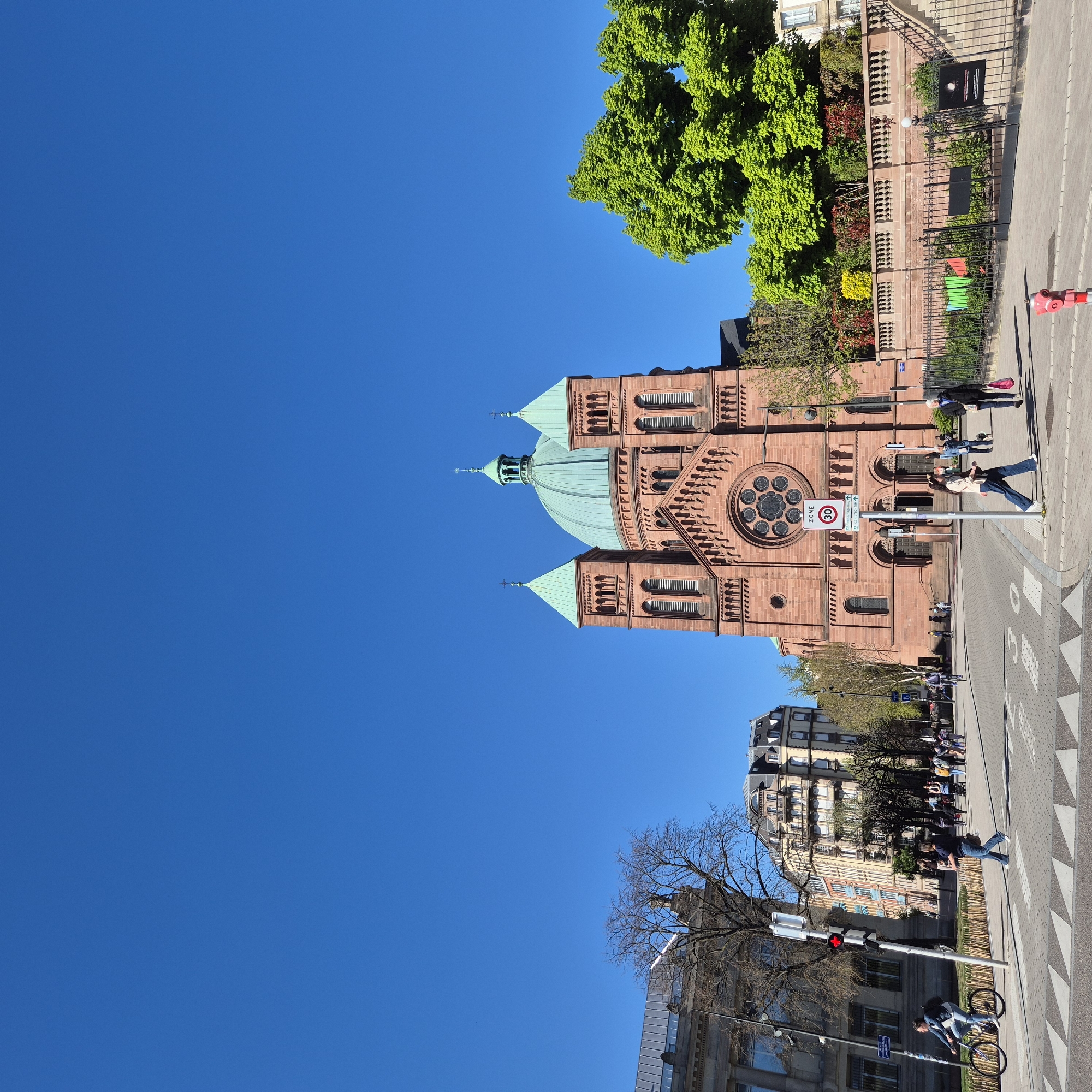 This image shows the Église Saint-Paul in Strasbourg, France. The perspective is from street level, looking upwards at the red brick church. Its green dome stands out against a clear blue sky. Part of the surrounding neighborhood is visible, including buildings, trees, sidewalks, and a road with pedestrians. The street includes traffic and pedestrian signals.