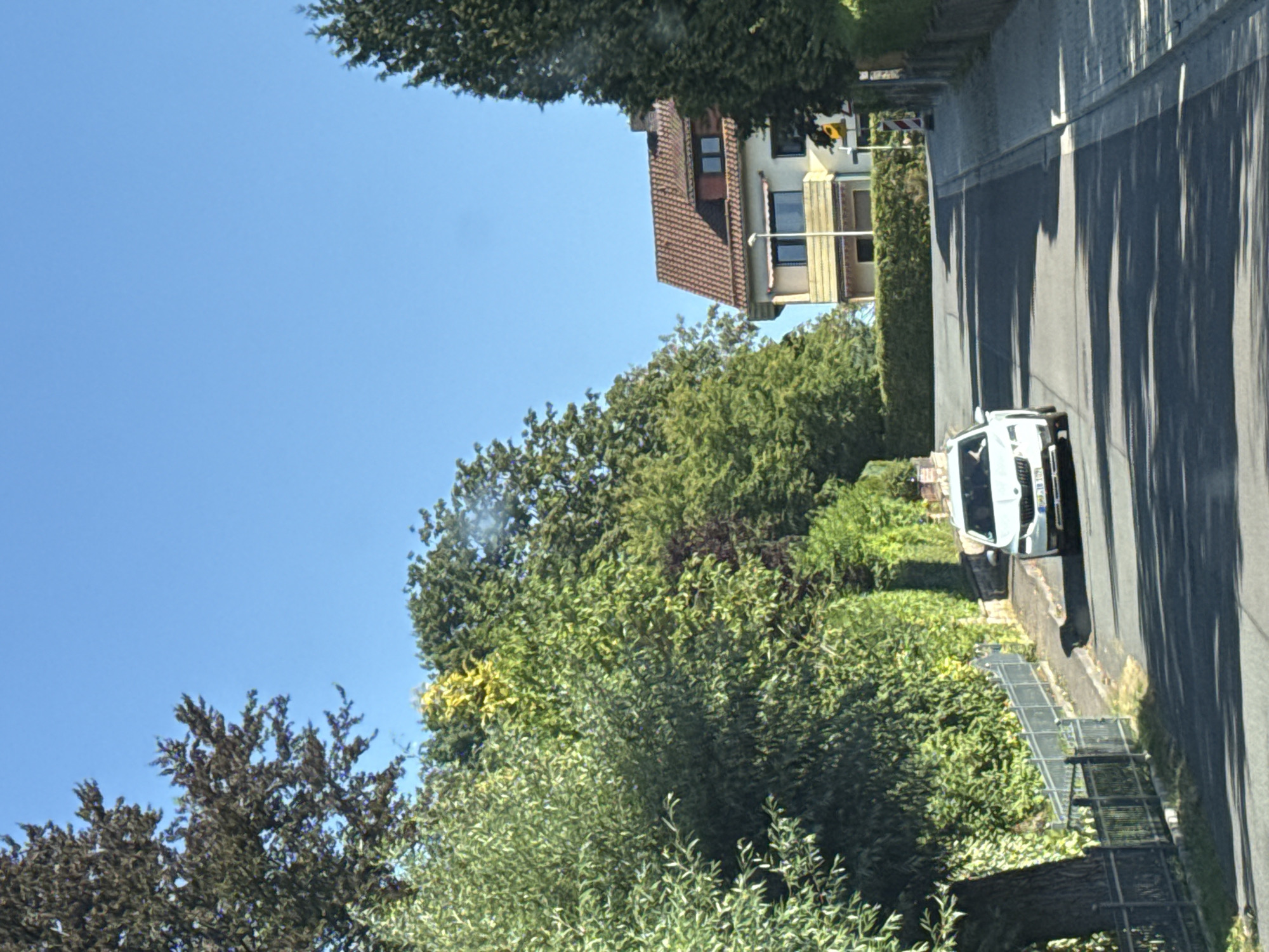 A white car is driving up a paved street lined with trees and houses in Bad Sachsa, Germany. The car has a license plate that reads "HRO IB 102". The street is bordered by a metal fence on the left and a sidewalk with stone paving on the right. A multi-story house with a red tile roof is visible among the trees on the right side of the street. The sky is clear and blue.