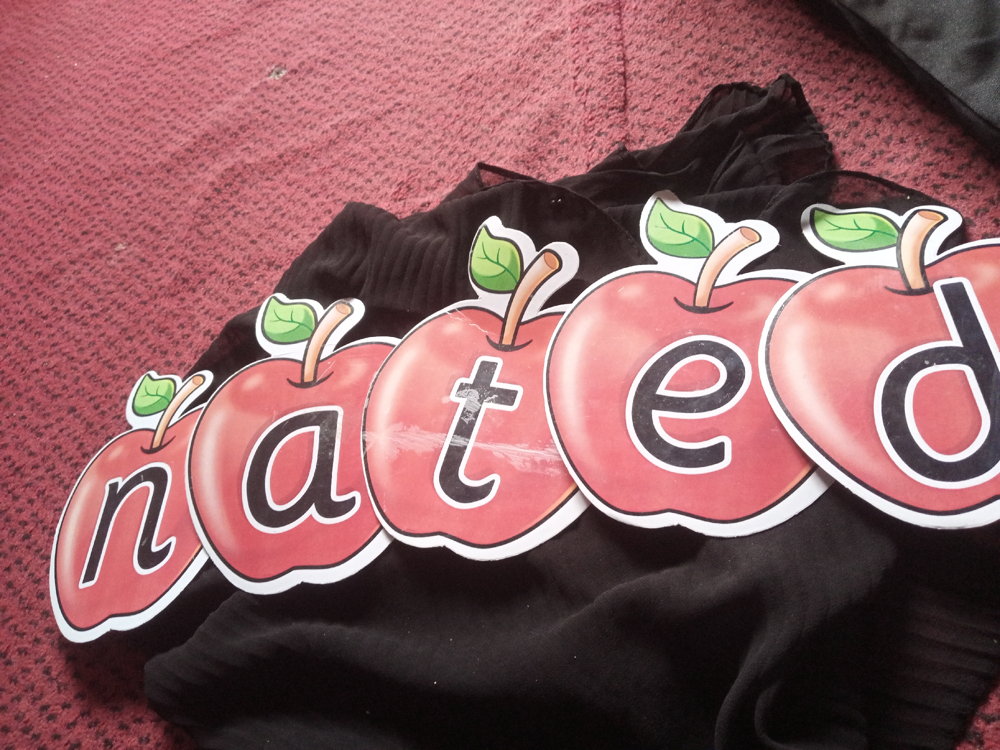 The image shows five apple-shaped cutouts, each featuring a letter. From left to right, the letters are "n", "a", "t", "e", and "d". The cutouts are arranged in a row and are placed on top of a black fabric. The background is a red, textured surface, possibly a carpet. The scene is indoors, and there are no people visible. The image appears to be a still photograph taken from a slightly elevated angle. There is no information in the image to suggest its location in Jalingo, Nigeria.