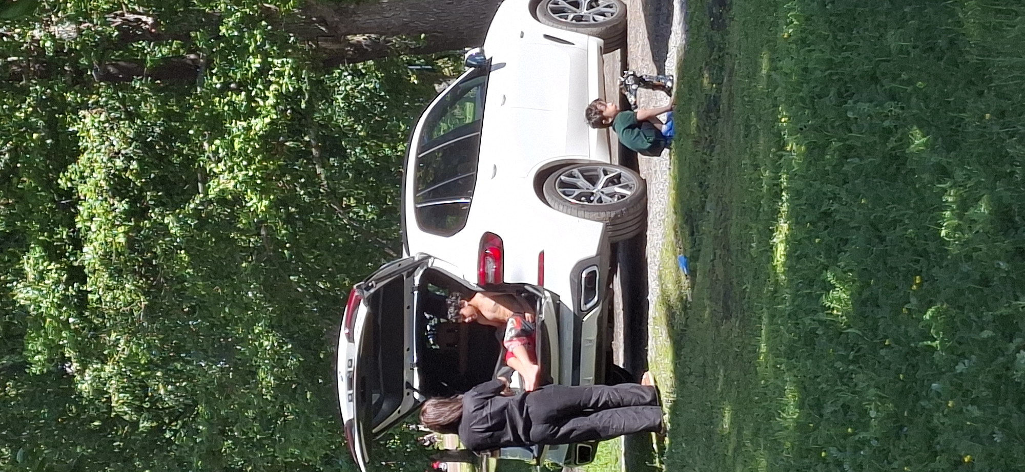 A white BMW SUV is parked on a gravel surface with its rear hatch open, revealing a young boy sitting inside. A woman stands near the open hatch, facing the boy. On the grass in front of the SUV, another young boy is playing with a toy. The scene is set outdoors with a grassy area and mature trees in the background, suggesting a park or wooded area. The lighting indicates it is daytime.