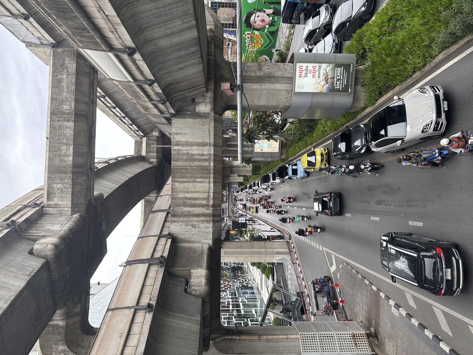 The image depicts a street scene in Bangkok, Thailand, characterized by heavy traffic and elevated infrastructure. The view is from an elevated position, looking down upon a busy road filled with cars and motorcycles. A concrete elevated train track structure dominates the left side of the frame. The road runs along the right side, with vehicles moving in the same direction. Advertisements and signs are positioned along the roadside. Buildings are visible along the edge of the road.