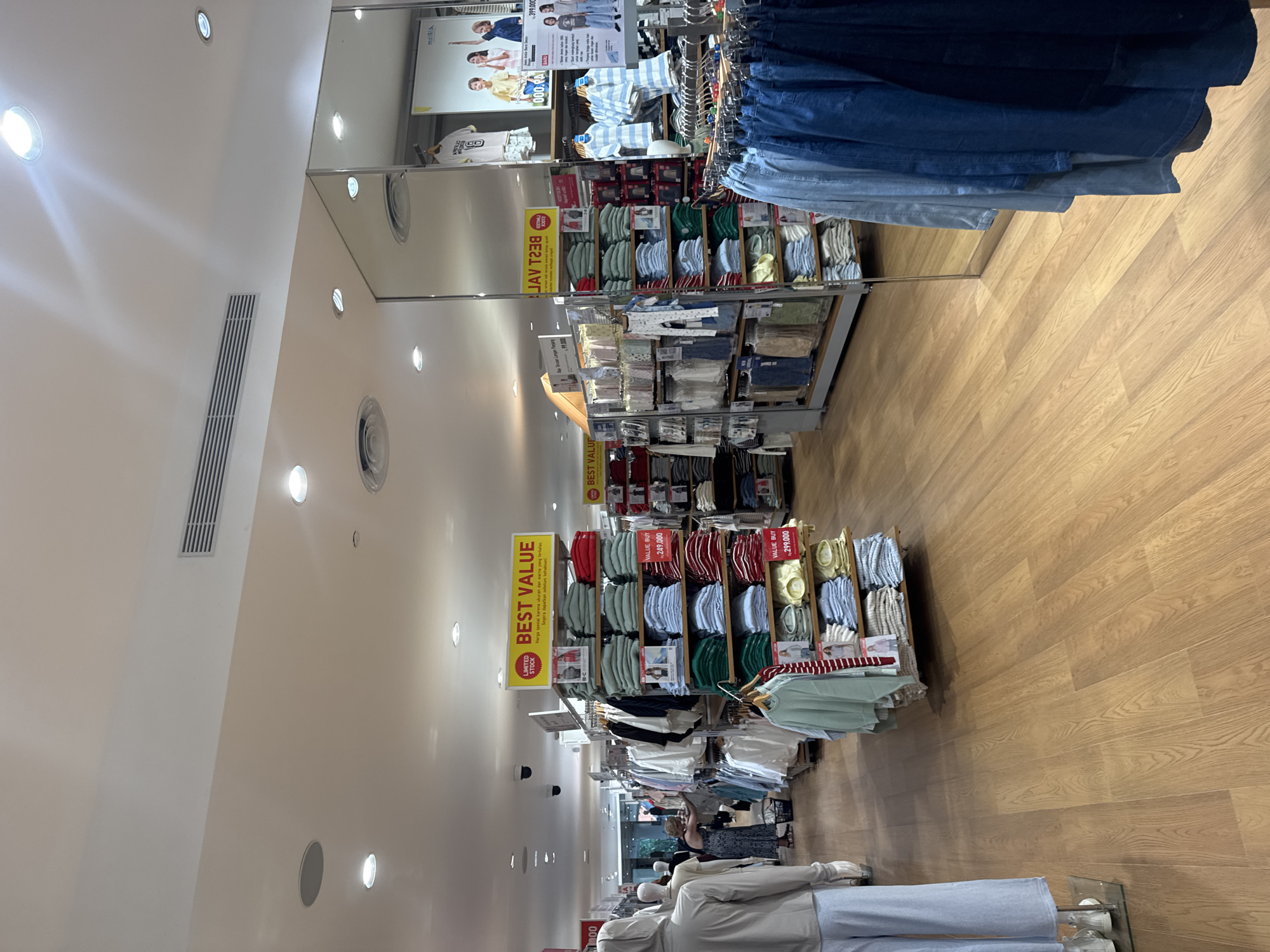 In a retail clothing store in Kuta, Indonesia, stacked shelves of folded t-shirts in various colors are displayed. Mannequins dressed in light-colored outfits stand to the left of the frame, while racks of clothing and additional merchandise are visible throughout the store. A woman in a dark patterned dress is browsing clothing on a rack. The store features a wooden floor and a reflective mirrored wall. A large "BEST VALUE" sign with promotional text is prominently displayed. The scene is illuminated by overhead lighting.