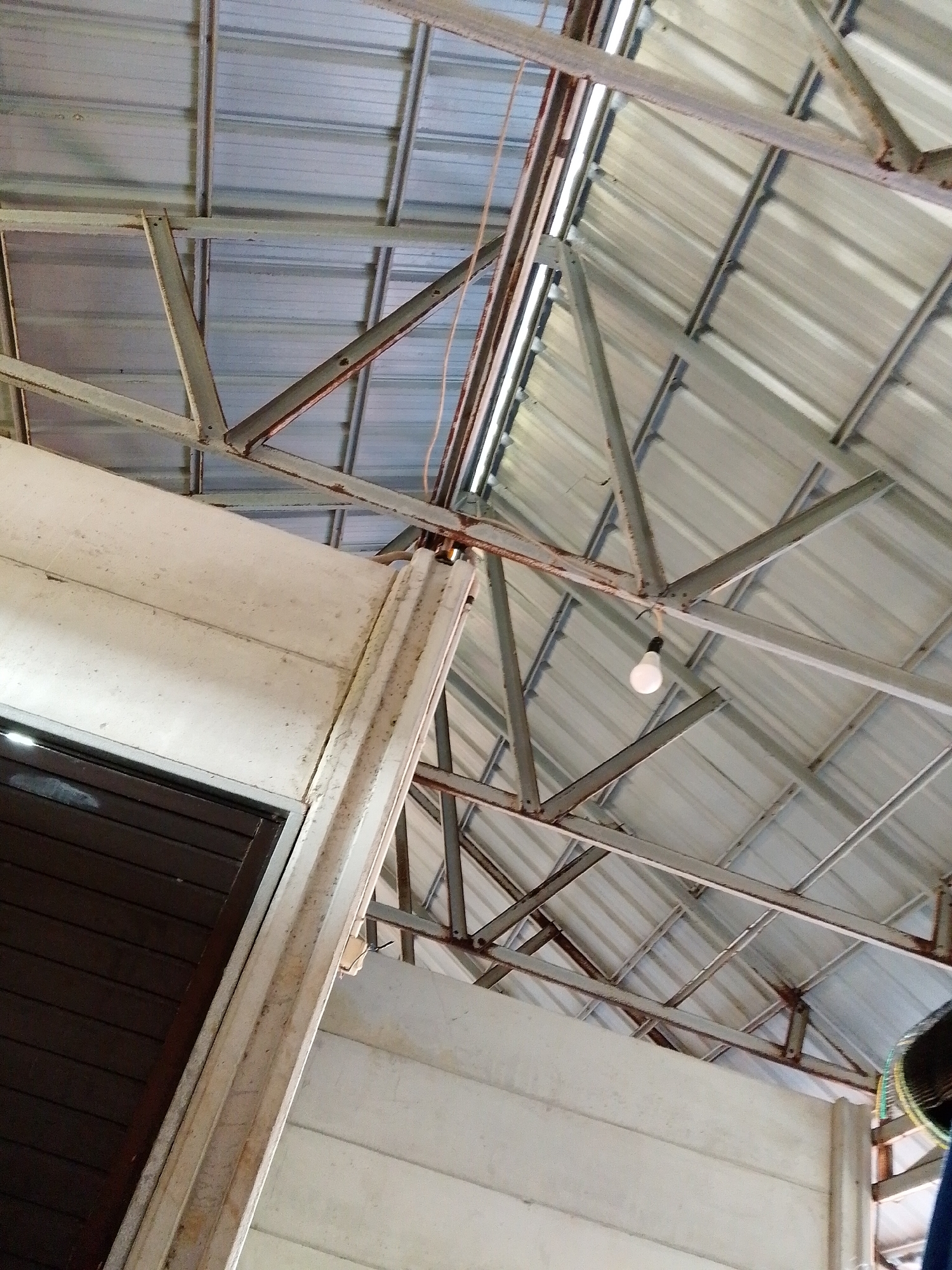 The image depicts the interior of a building in Bukit Ketok, Indonesia, specifically the ceiling and upper portions of the walls. The roof is made of corrugated metal sheeting supported by a network of steel beams and trusses. A single light bulb hangs from the roof's frame. The walls appear to be constructed from precast concrete panels, some showing signs of staining or weathering. A dark-colored door is visible along the left side of the frame.