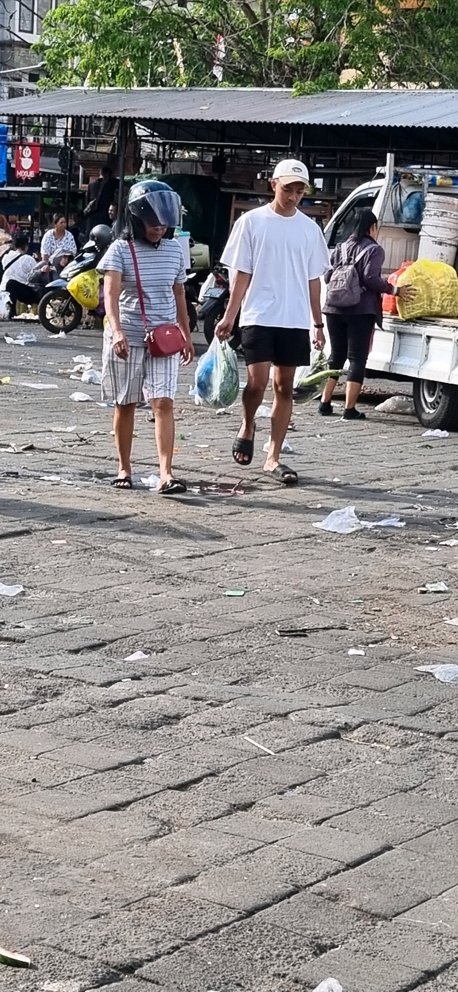 This is a medium shot, eye-level, outdoor vertical photograph taken in Denpasar, Indonesia. The scene depicts a market area with cobblestone paving littered with trash and debris. In the foreground, two people are walking away from the camera. The person on the left, identified as a female, is wearing a blue and white striped t-shirt, plaid shorts, black flip-flops, and a black helmet. She is carrying a red purse over her right shoulder. The person on the right, identified as a male, is wearing a white t-shirt, black shorts, and black flip-flops. He is carrying a clear plastic bag containing produce, possibly a watermelon, in his right hand. Behind them, a white truck is parked, loaded with large yellow bags of goods. Other individuals and a parked motorcycle are visible in the background, suggesting a busy marketplace environment. Trees and market stalls with corrugated metal roofs are also present. The lighting suggests it is daytime, possibly late morning or early afternoon, given the shadows.