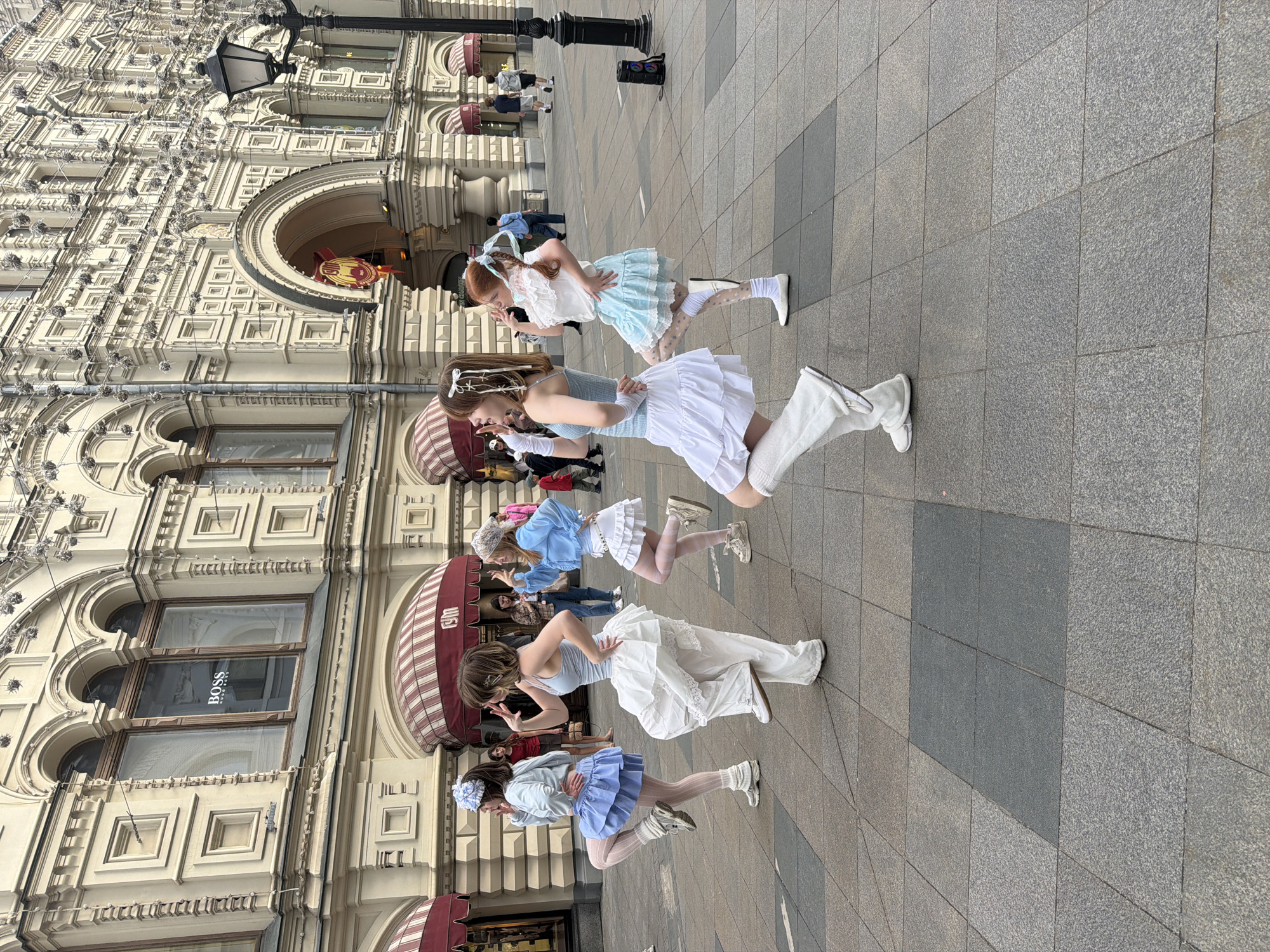 A medium-angle outdoor shot presents a group of five women in an open public space in front of a grand building. The women are arranged in a line, posed in a synchronized manner with one leg extended forward and arms positioned in front of them. They are dressed in coordinated pastel-colored outfits consisting of short skirts, tops, tights, and footwear, with some wearing knee-high socks or boots.
The setting appears to be a paved public square in Moscow, Russia, as evidenced by the GUM department store in the background. The building's facade is ornate, with arched windows, architectural detailing, and striped awnings above storefronts. A "BOSS" sign is visible in one of the windows. Pedestrians and a street light fixture are also visible in the background. The pavement is composed of large square tiles in varying shades of gray.