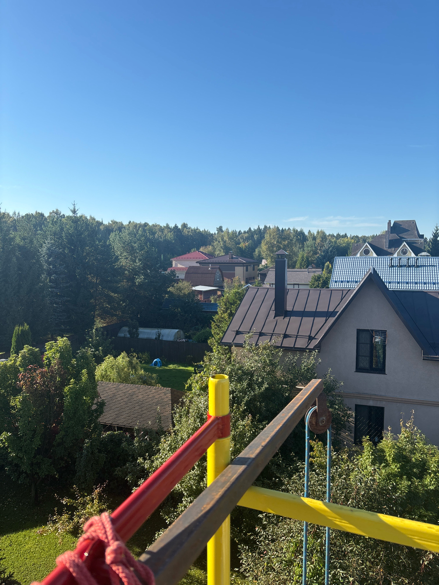 The image is a high-angle view of a suburban residential area with numerous houses and dense forestation under a clear blue sky. In the foreground, a section of a metal structure, painted yellow and red with a rope attached, is visible, suggesting it might be part of a playground or observation point.
The midground features several houses with diverse roof styles, including dark brown, light blue tiled, and gray pitched roofs. One prominent house has a dark brown metal roof with a black chimney. Other houses are partially obscured by trees. A greenhouse or poly tunnel is visible in the middle-left area, and a small blue inflatable object is on a grassy lawn.
The background is dominated by a thick, green forest. The overall scene suggests a tranquil, possibly rural or semi-rural setting within the city of Zhavoronki, Russia. The lighting indicates it is daytime, likely late morning or early afternoon, with bright sunlight casting shadows. No people are directly visible in the frame.