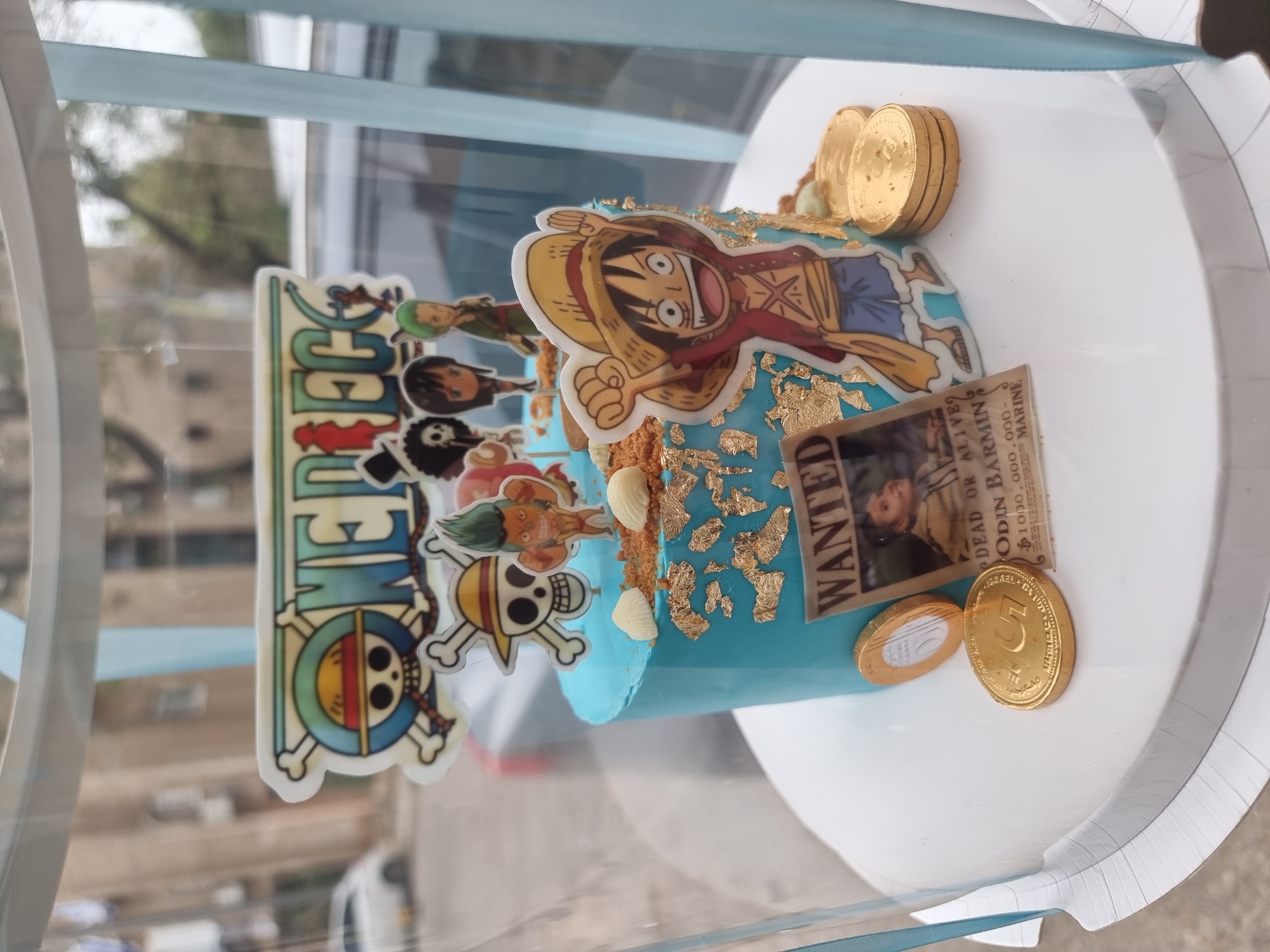 The image shows a One Piece-themed cake inside a clear plastic box. The cake is cylindrical with light blue frosting decorated with gold leaf, white chocolate pieces, a One Piece character topper, and a "wanted" poster replica. Stacks of gold chocolate coins are arranged on top and around the cake. The cake sits on a white circular base. The plastic box has a light blue trim. Outside the box, blurred backgrounds suggest an urban environment, possibly in Tel-Aviv, Israel, with buildings and vegetation visible.