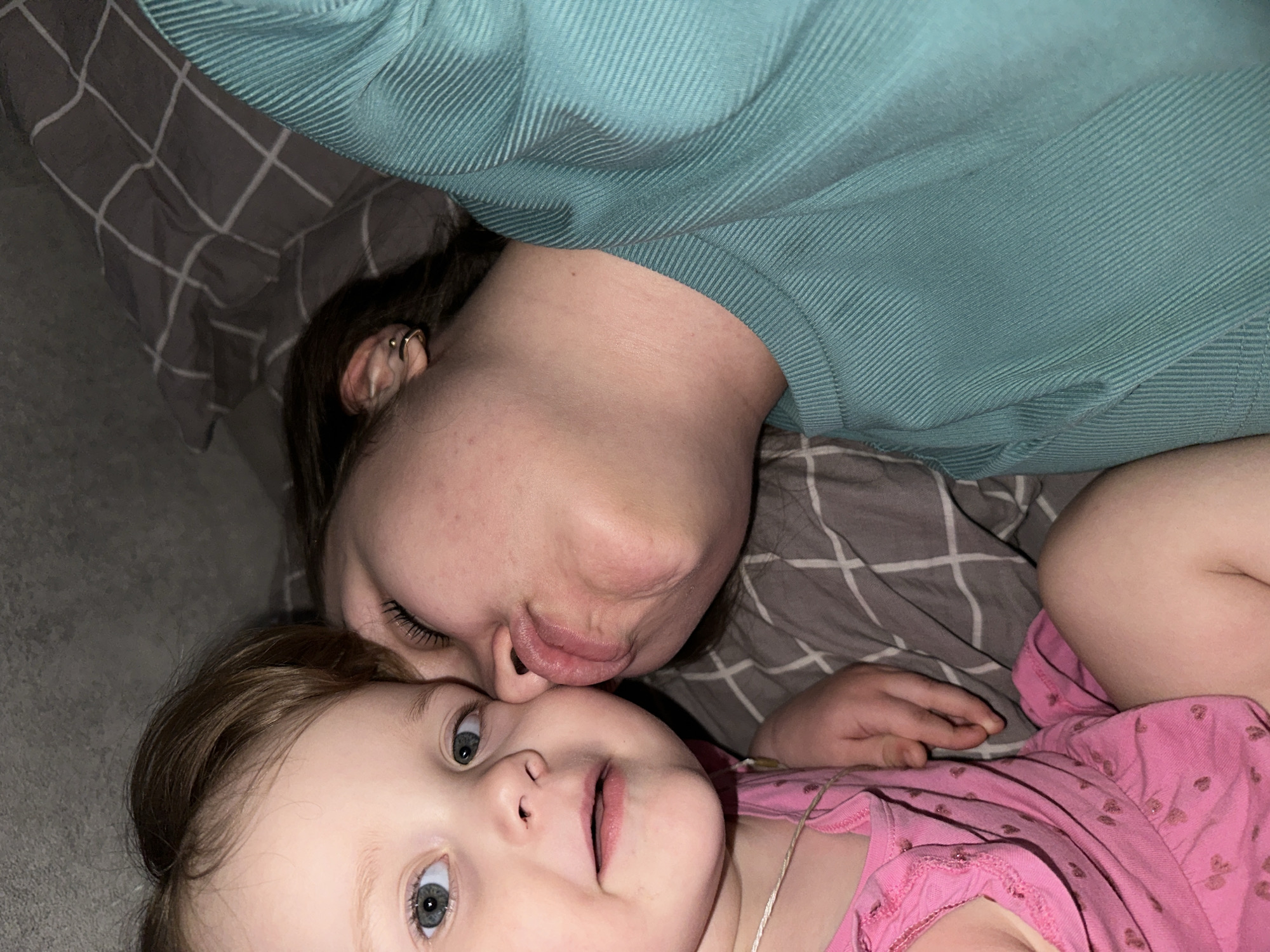 A woman and a young child lie on a bed covered with a gray and white checkered blanket. The woman is wearing a turquoise ribbed shirt, and the child is wearing a pink shirt with small heart patterns. The woman is positioned above the child, with her face close to the child's face, appearing to kiss or nuzzle her. The child is looking at the camera with a neutral expression. The setting appears to be indoors, possibly a home. There is no information to identify the location specifically as Chaikovsky, Russia.