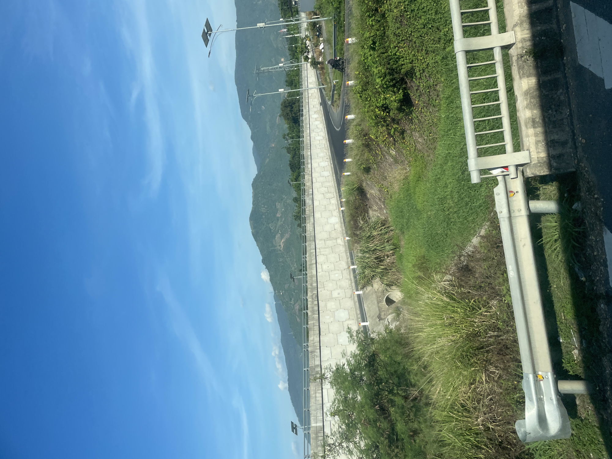 The image shows a section of a road in Tay Nha Trang Ward, Vietnam, under a clear blue sky with scattered clouds. A multi-level road structure dominates the scene, with a concrete overpass or retaining wall on the left, featuring a textured surface. The road curves to the right, with solar-powered streetlights lining its edge. Lush green vegetation, including tall grasses and trees, grows on either side of the road and in the lower foreground. A concrete culvert is visible in the grassy area. A motorcycle with a rider is seen on the road, moving towards the right side of the frame. In the background, rolling hills covered in trees rise against the horizon. A metal guardrail with vertical slats is present in the immediate foreground, partially obscuring the view of the road and vegetation.
