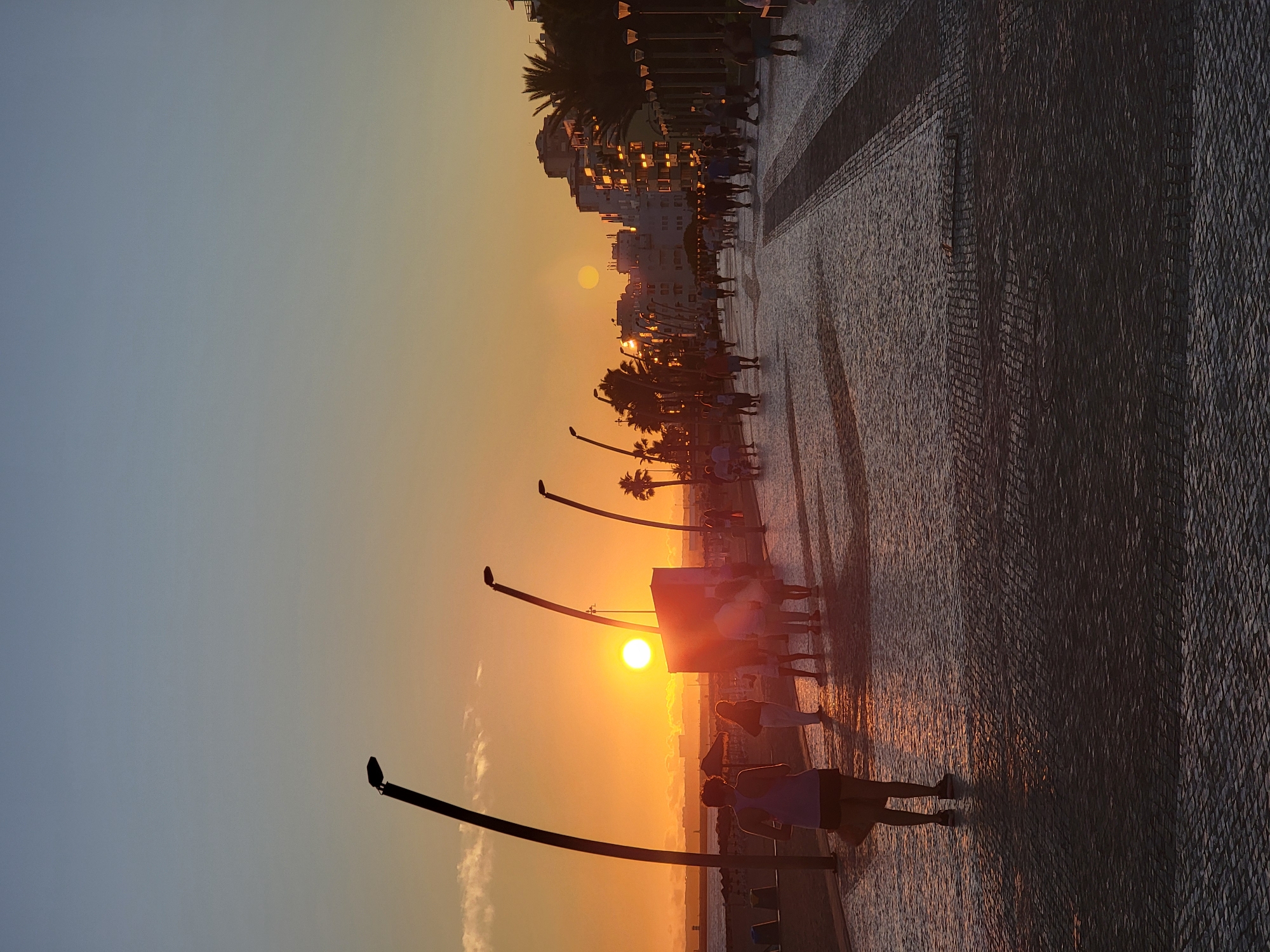 A vertical, medium-shot photograph captures a sunset scene on the Quarteira promenade in Quarteira, Portugal. The sun, a bright orange orb, is low in the sky, casting a warm glow over the scene. Silhouetted figures of people stroll along the wide, paved promenade, their shadows stretching long across the cobblestone walkway. A lone figure in the foreground, viewed from behind, is wearing a purple tank top and dark shorts, with a bag slung over their shoulder. In the mid-ground, several groups of people are walking, some in conversation. The promenade is lined with decorative lamp posts, their dark, slender frames creating a repeating pattern. Beyond the promenade, the ocean is visible, with a few distant buildings and palm trees lining the coastline. The buildings appear to be hotels or apartments, illuminated by the setting sun. The sky transitions from a pale blue at the top to a soft orange and yellow hue near the horizon. The overall atmosphere is one of peaceful evening activity, with the natural beauty of the sunset enhancing the urban setting.