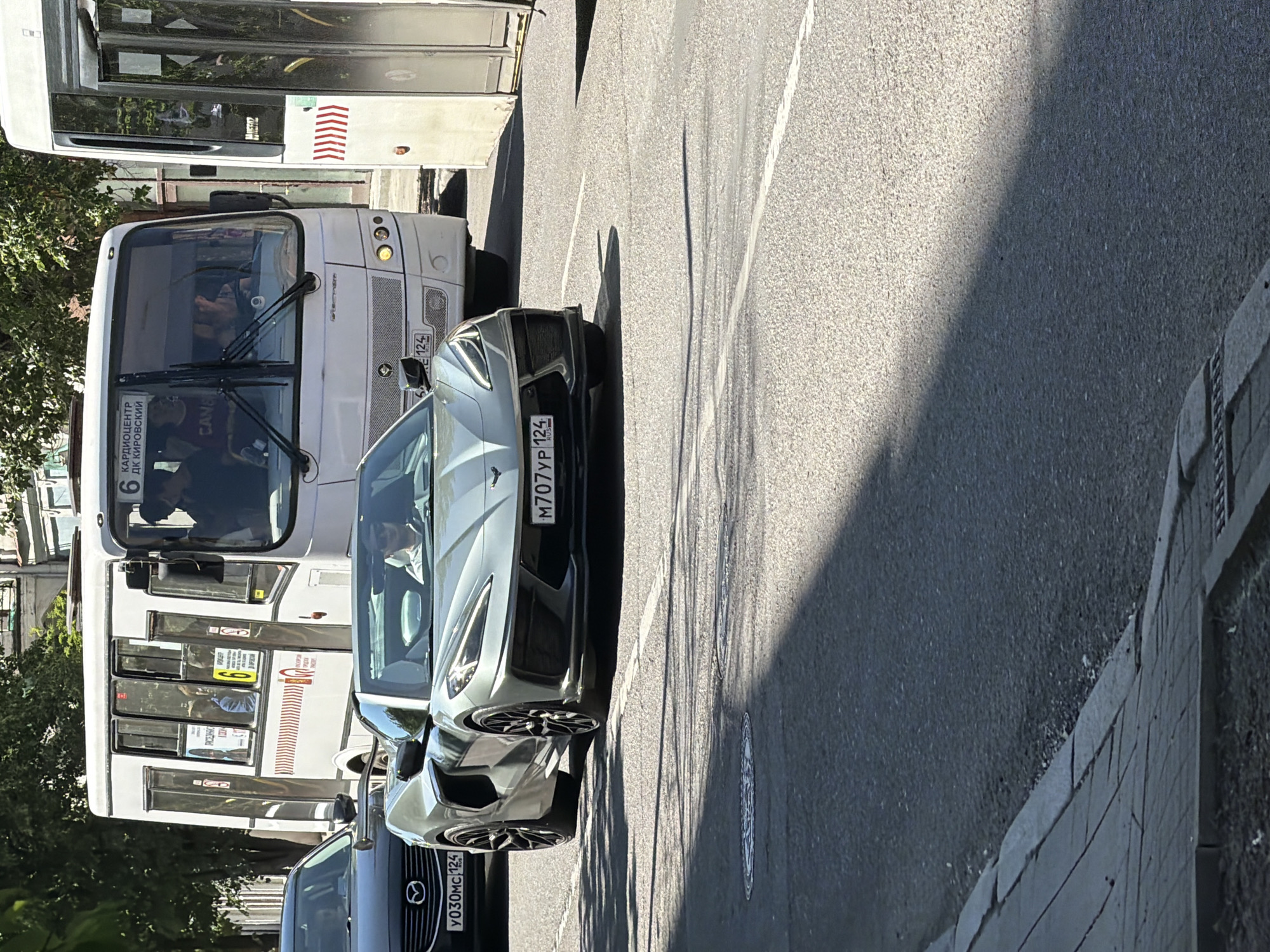 This media file captures an outdoor scene on a sunny day, likely during daylight hours. The primary subjects are vehicles, including a white bus and two cars, positioned on a paved road. The bus, on the left, appears to be a public transport vehicle with visible signage in Russian. One car, a light gray sports car, is parked next to the bus, and another, a dark sedan, is partially visible in the foreground. The scene is set on a street with a sidewalk made of stone and asphalt. The time of day is suggested by the shadows cast by the vehicles and the bright sunlight. No specific activities or events are clearly depicted, beyond vehicles being parked or stopped on a street.
Visible text includes Russian inscriptions on the bus and license plates on the cars. The bus has "КАРДИОЦЕНТР ДК КИРОВСКИЙ" (Cardiology Center, Kirovsky District Palace of Culture) and the number "6" displayed. The license plates on the cars are "M707YP 124 RUS" and "YO30MC 124". The location context indicates Krasnoyarsk, Russia. The overall mood is neutral, focusing on the urban streetscape and vehicles.