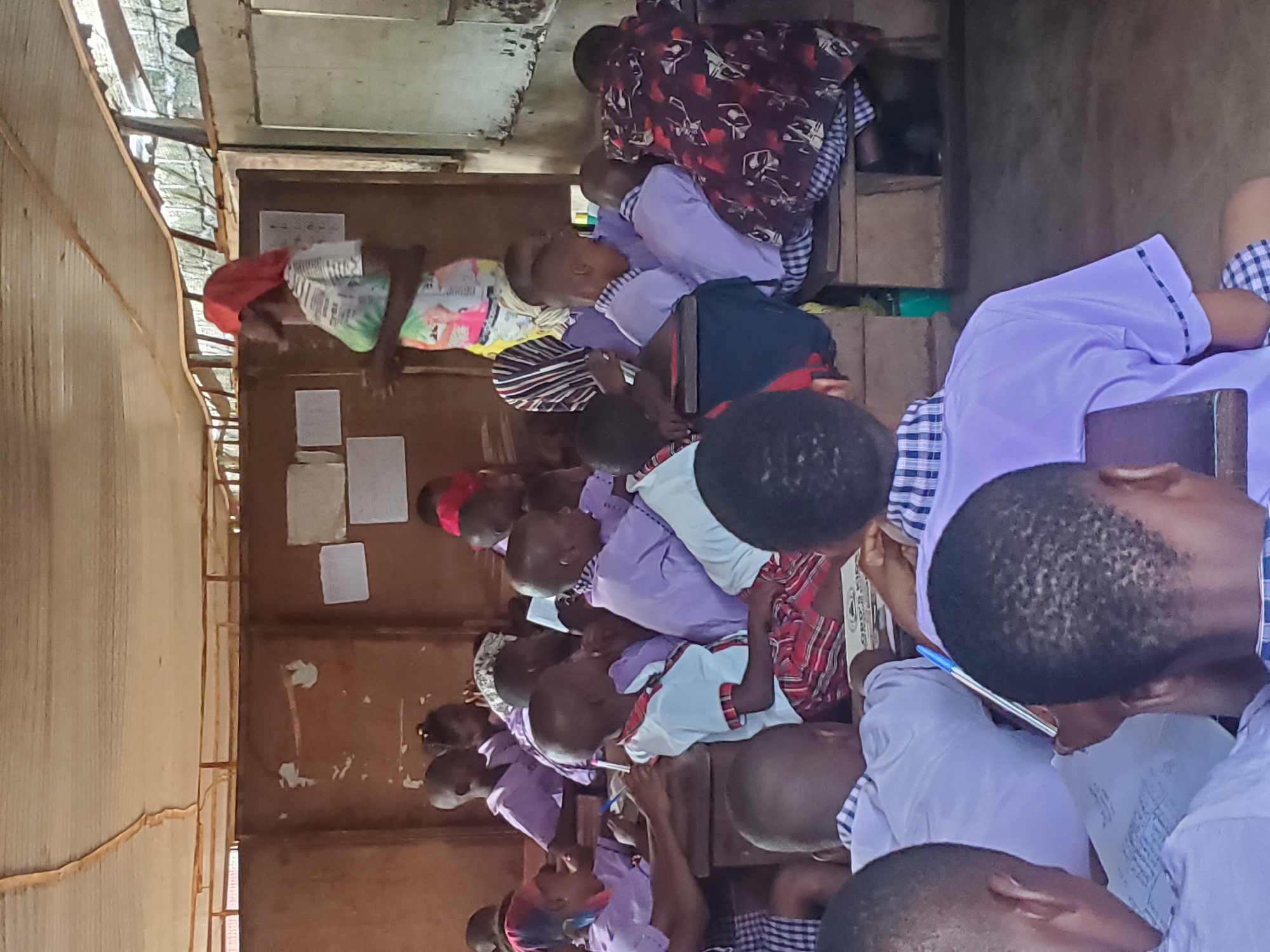 The image shows a classroom in Ukwu-Abwa, Nigeria. Students are seated at desks, some writing. A woman in a red headscarf stands in the corner. The classroom has a corrugated metal wall, and there are papers on the wall. Another adult sits in the back, and appears to be reading something.