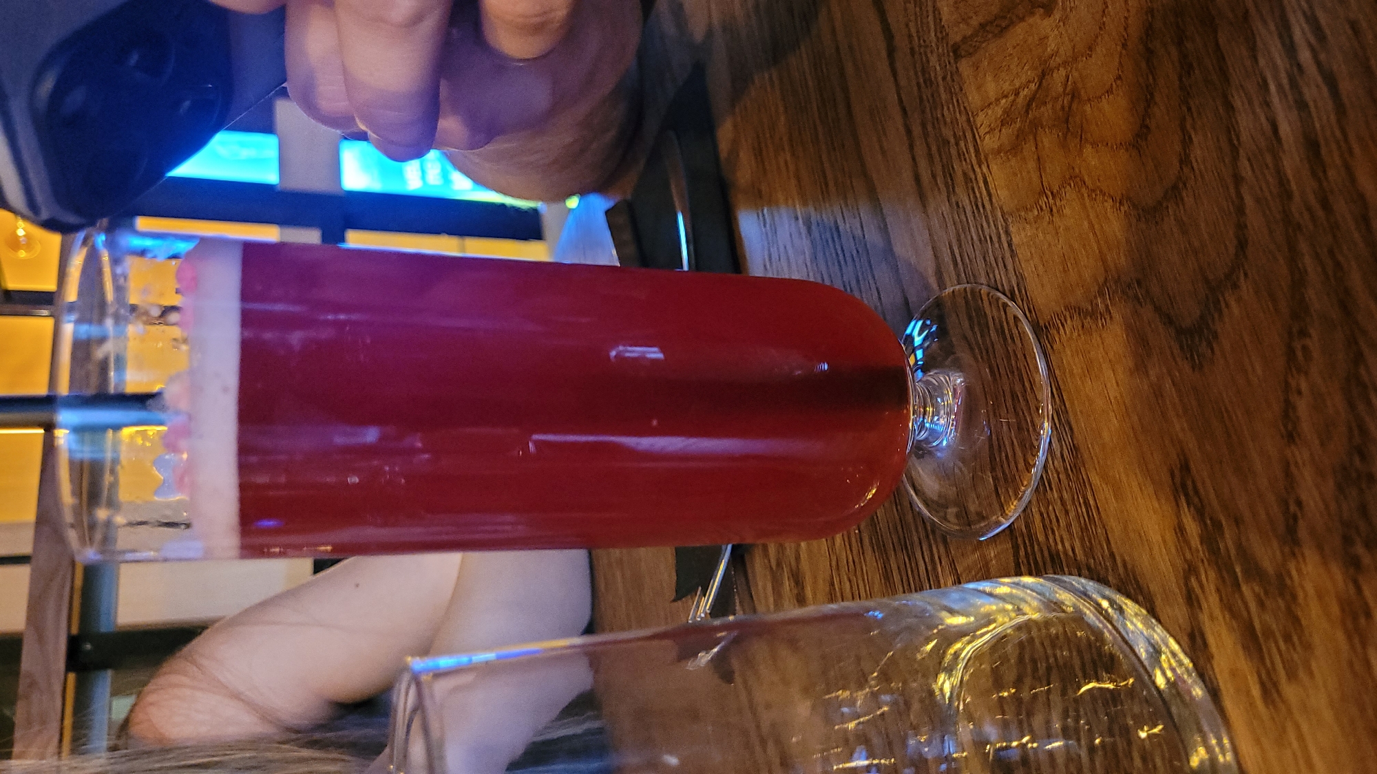 In Sofia, Bulgaria, at a dimly lit establishment with wooden furniture, a tall glass of red, frothy beverage sits on a wooden table. A person's arm, partially visible on the left, is close to an empty glass. On the right, a hand holds a dark-colored smartphone, its screen illuminated. The background features a decorative wall with yellow and blue elements. The beverage appears to be a cocktail or a juice.