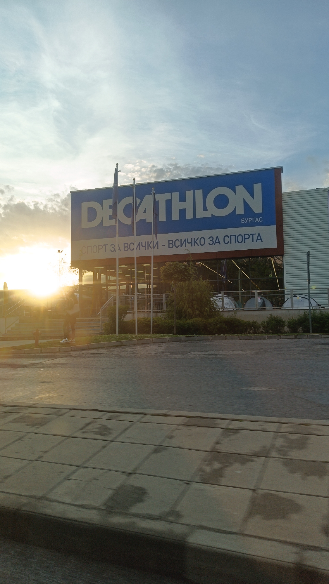 The image shows the Decathlon store in Burgas, Bulgaria. The store is identifiable by its large blue and white Decathlon sign, which also includes the city name in Bulgarian (БУРГАС). Below the store's name is the slogan "СПОРТ ЗА ВСИЧКИ - ВСИЧКО ЗА СПОРТА", which translates to "Sport for all - everything for sport." The store has a grey facade with vertical lines. Several flagpoles stand in front of the store.
A man is standing near the entrance, with a bright sun behind him. In the background, through the store's glass, several tents are visible.
In the foreground is a wet pavement, suggesting it recently rained. The road in front of the store is visible. The image seems to have been taken from a slightly elevated position, possibly from a vehicle.